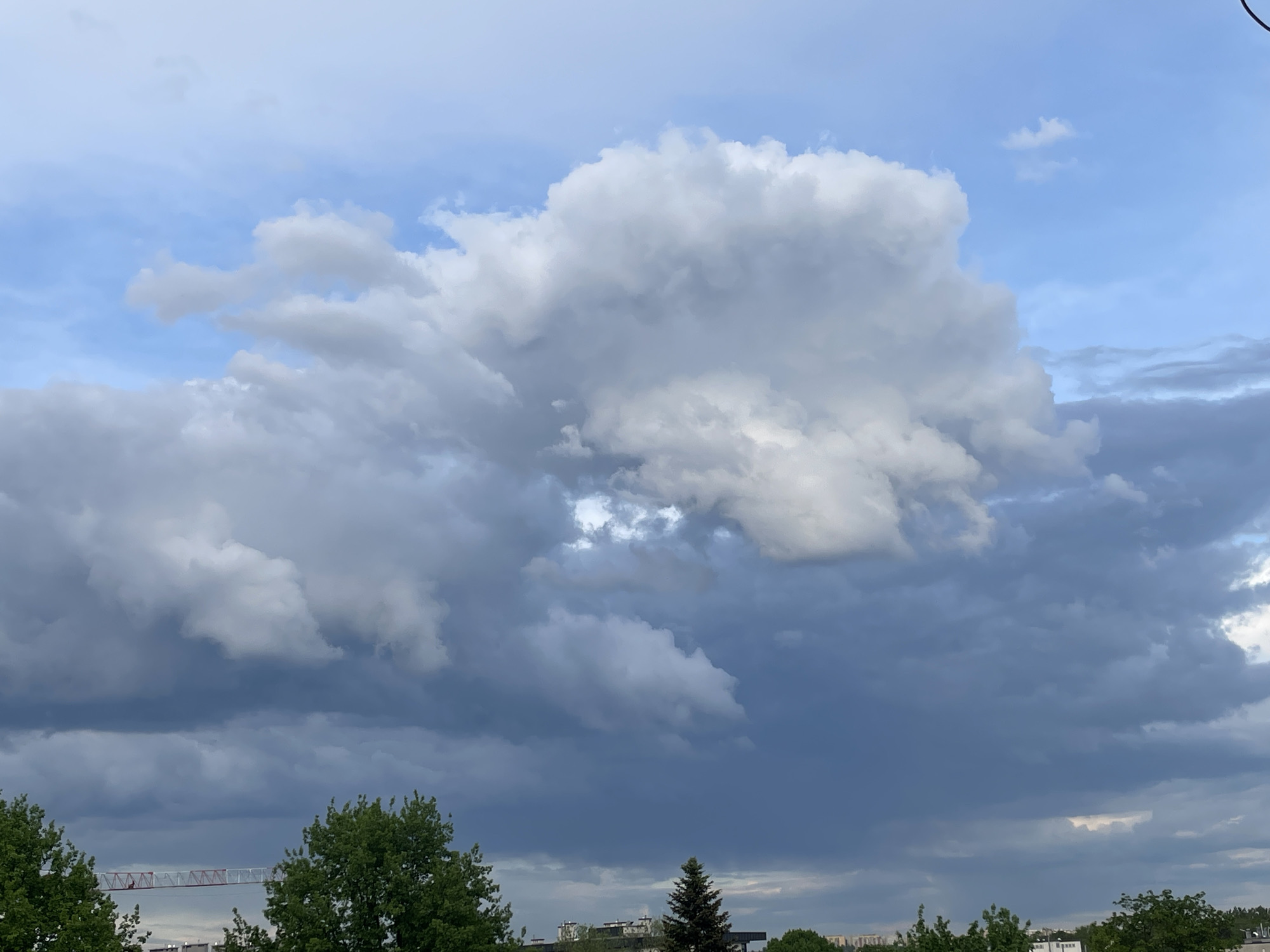 The image depicts a cloudy sky over what appears to be a cityscape. Dominating the view is a large, cumulonimbus cloud formation with varying shades of gray and white, suggesting possible rain. Blue sky is visible in some areas between the clouds. At the bottom of the image, there are trees and glimpses of buildings, indicating an urban setting. A construction crane is also visible among the trees. The exact location within the city of Unknown, Unknown is not discernible from the image.
