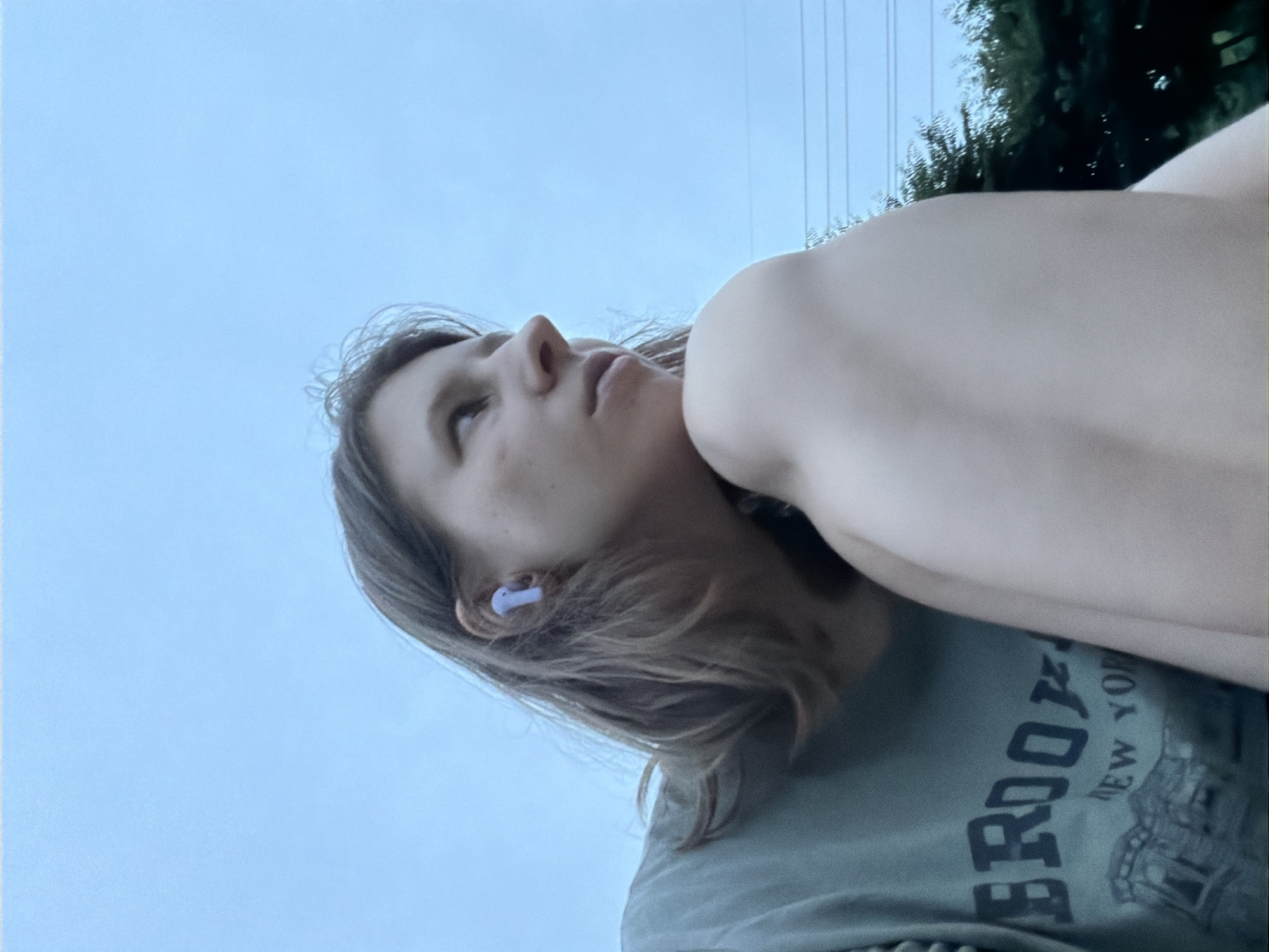 In an outdoor setting, a young woman with light brown hair is captured from the chest up, looking to her right. She is wearing a teal t-shirt with the word "BROOKLYN" and "NEW YORK" printed on it in a faded, dark font, with a graphic of a cityscape below. A white earbud is visible in her left ear. Her right arm is bent at the elbow, with her hand resting near her chin. The background consists of a pale blue sky with a few thin, dark horizontal lines that could be power lines, and dark green trees are visible in the lower right corner. The lighting suggests it might be dusk or an overcast day. The image is a close-up, low-angle shot. The location is specified as Dongducheon-si, South Korea, although no specific landmarks within the city are visible.