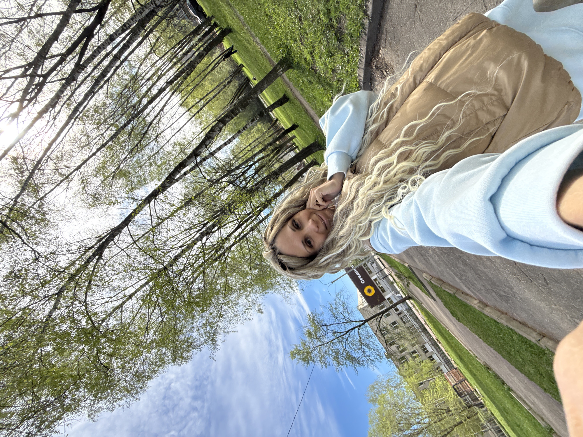 A woman with long blonde hair is taking a selfie. She is wearing a light blue sweatshirt and a tan puffy vest. The background shows trees with green leaves, a blue sky with white clouds, and a building with a sign that includes a sunflower and the word "СРЕДНЕ". This photograph appears to have been taken in Rusanovo, Russia.