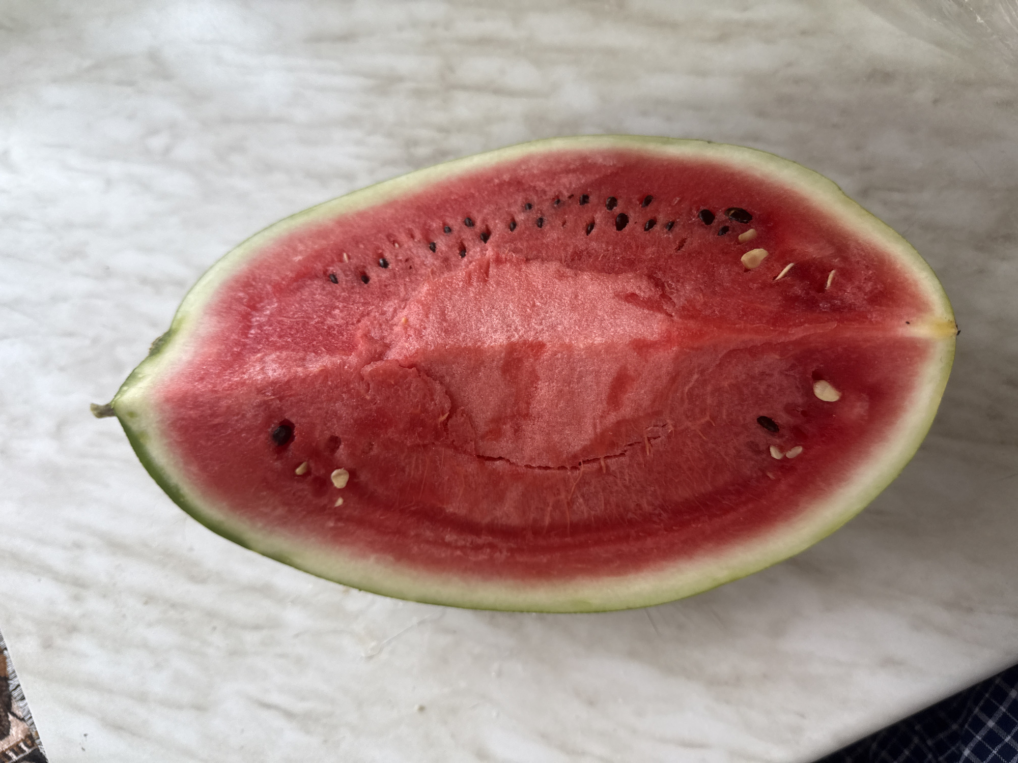 A top-down, eye-level shot shows a slice of watermelon on a white, marble-like surface. The watermelon is cut in half, revealing its bright pink flesh and a few scattered seeds. The rind is a light green color. The background is mostly out of focus, but a portion of a dark blue and white checkered cloth is visible in the lower left corner. The lighting is natural and appears to be from an overhead source. The overall impression is one of freshness and simplicity.