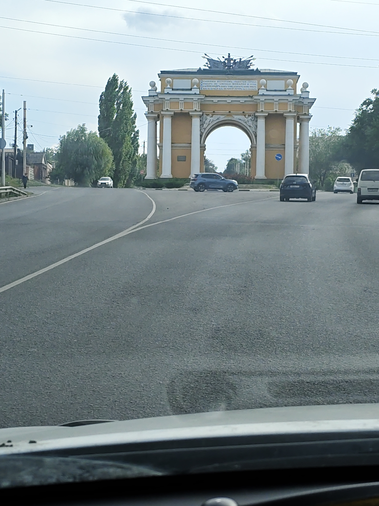 The image shows the Novocherkassk Arch of Triumph in Novocherkassk, Russia. The arch is a large, yellow structure with white columns and decorative elements. The inscription on the arch is visible. In front of the arch, there is a road with several cars driving on it. A blue SUV is in the left lane, a dark SUV is in the right lane, and two white cars are further ahead. The road is flanked by trees and buildings on the left side. The sky is cloudy. The perspective suggests the photo was taken from inside a vehicle, looking forward.