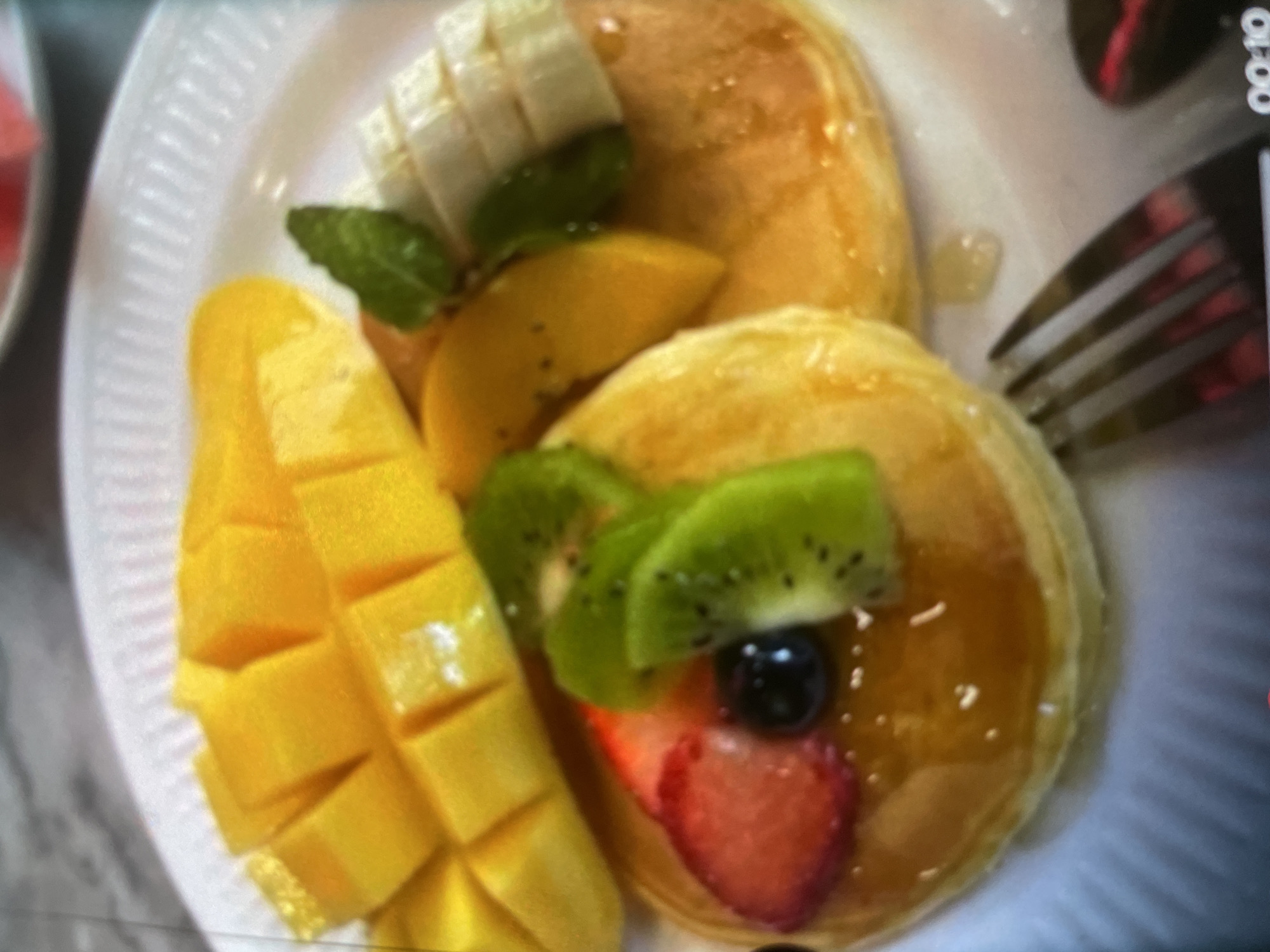 A close-up shot shows a plate of pancakes adorned with fresh fruit. The pancakes are drizzled with a golden syrup and topped with sliced strawberries, kiwi, and blueberries. A few slices of mango are arranged on the plate, alongside some sliced bananas and a sprig of mint. The plate itself is white and appears to be made of a disposable material. A fork is visible on the lower right side of the image, hinting at a meal about to be enjoyed. The background is blurred, but suggests a dining setting. No specific location within Turkistan, Kazakhstan can be identified from the image.