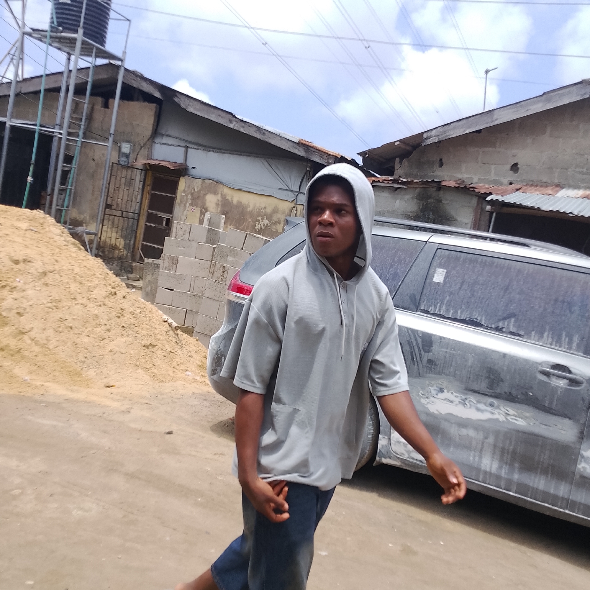 In Ado, Nigeria, a young Black male wearing a grey hooded sweatshirt and dark blue jeans walks past a dusty, silver SUV. He is captured from the chest up, with his head turned to the right, looking away from the camera. The environment appears to be an outdoor setting with a pile of sand to the left, and buildings constructed from concrete blocks and corrugated metal roofing to the right. Electrical wires are visible overhead against a cloudy sky.