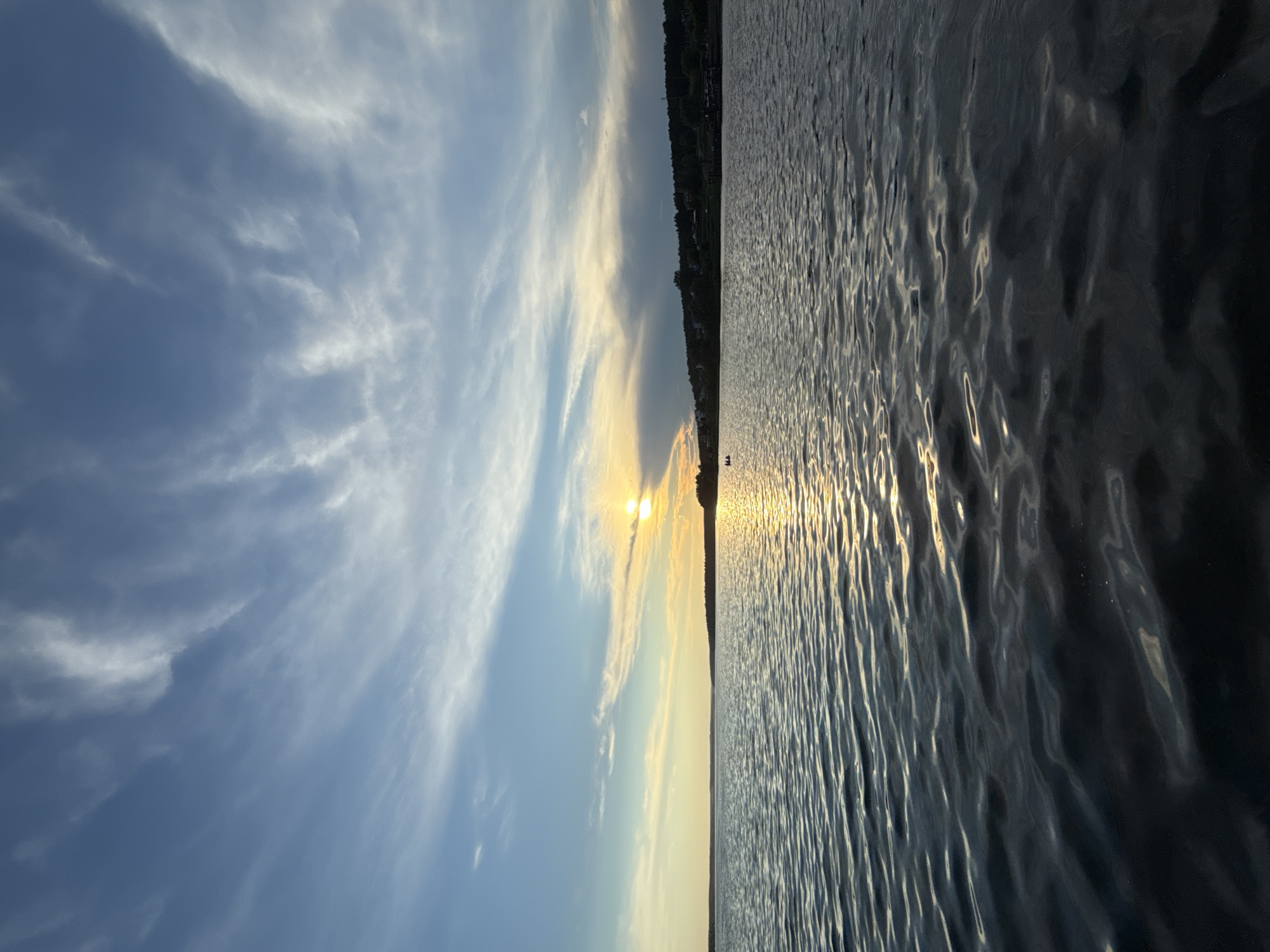 This is a vertical, wide-angle photograph taken at eye-level, depicting a serene lake at sunset in Votkinsk, Russia. The sky dominates the upper half of the image, displaying a gradient of blue, transitioning from a pale azure near the horizon to a deeper blue overhead. Wispy, white clouds streak across the sky, catching the last rays of the setting sun. The sun itself is partially obscured by clouds, casting a warm, golden light and creating a shimmering reflection on the water's surface.
The lower half of the image features the calm, rippling surface of a lake. The water is a deep blue-gray, reflecting the sky and the golden hues of the sunset. Gentle waves create a textured surface, with highlights of light dancing across them.
In the distance, along the horizon, a dark, forested shoreline is visible, punctuated by a few scattered houses and buildings. The trees appear dense and dark against the brighter sky. Two small figures in a boat are visible in the far distance, to the left of the sun's reflection on the water, appearing as small dark silhouettes against the bright water. On the right side of the shoreline, a larger boat is docked. The overall atmosphere of the image is tranquil and peaceful, characteristic of a quiet evening by the water.
