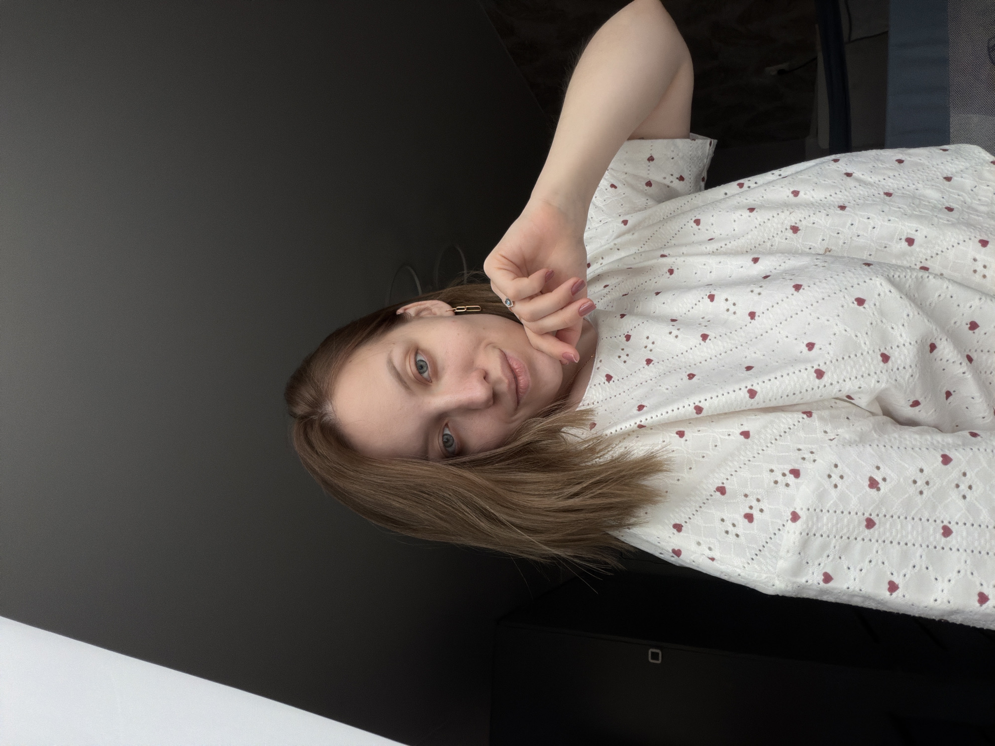 An image, originally oriented vertically but showing a horizontally composed scene, features a young woman lying down and looking towards the camera. She has light brown hair, which falls around her face, and blue eyes. Her expression is calm and appears to be a slight, soft smile. She is wearing a white top or dress adorned with delicate eyelet lace patterns and small red hearts. Notable accessories include a minimalist gold-toned earring shaped like a paperclip in her left ear and a ring on one of her fingers. Her fingernails are painted with light pink polish.
The woman occupies the right side of the frame, with her head towards the top and her body extending downwards. Her left arm is bent, and her hand is resting near her face.
The background is predominantly dark grey to black, suggesting an indoor setting. There's a subtle distinction in the background where a lighter grey area meets a darker, almost black, section, possibly indicating a wall meeting a piece of furniture or a darker floor. The overall lighting is soft, characteristic of indoor conditions, without strong shadows or bright natural light sources visible. There is no visible text in the image. The scene takes place in Reshetnikovo, Russia.