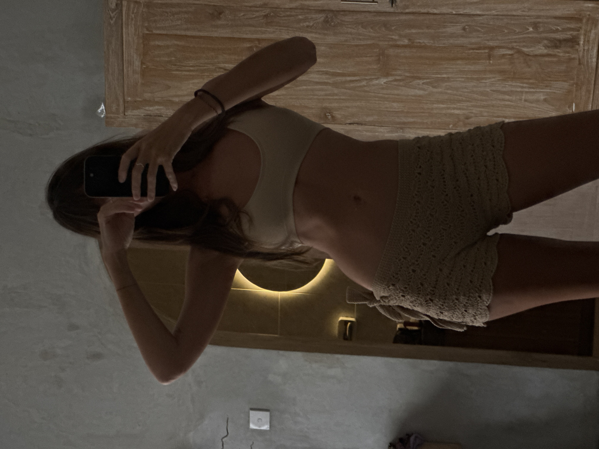A woman is taking a selfie in a bathroom mirror in what appears to be North Kuta, Indonesia. She is wearing a light-colored sports bra and crochet shorts. A vanity and a lit circular mirror are behind her. The bathroom has grey concrete walls and dark brown doors and trim.