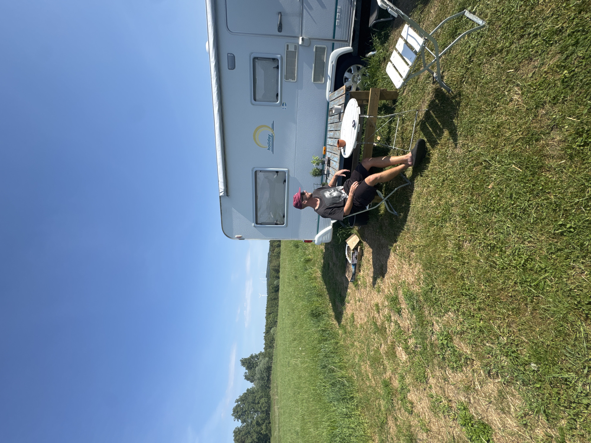 The image depicts a bright, sunny outdoor scene featuring a campervan parked on a grassy field. The sky is a clear, cloudless blue, suggesting pleasant weather, likely midday or early afternoon given the strong sunlight and shadows.
A person, likely an adult male, is seated on a white folding chair next to the campervan, facing slightly towards it. He wears a dark t-shirt, dark shorts, and a red baseball cap, appearing relaxed with one bare foot and one foot in a dark slipper. Beside him is a small wooden table with a white circular top, holding an orange mug and some small potted plants. Another empty white folding chair is visible nearby.
The campervan is light-colored (white/light grey) with a green/blue stripe running along its side. It features windows, a door, and a partially visible tire. On its side, the word "holiday" is visible, accompanied by a stylized sun logo. Faint numbers, possibly "353", are also present higher up on the vehicle. Some miscellaneous items in a box are near the campervan's front wheel.
Beyond the campervan and foreground, a vast expanse of green field stretches towards a distant treeline. In the far background, above the trees, several wind turbines are visible, indicating a rural or semi-rural setting. The ground is primarily green grass, with some drier, brownish patches. The scene conveys a sense of leisure and outdoor relaxation, likely during a camping trip in Vessigebro, Sweden.
