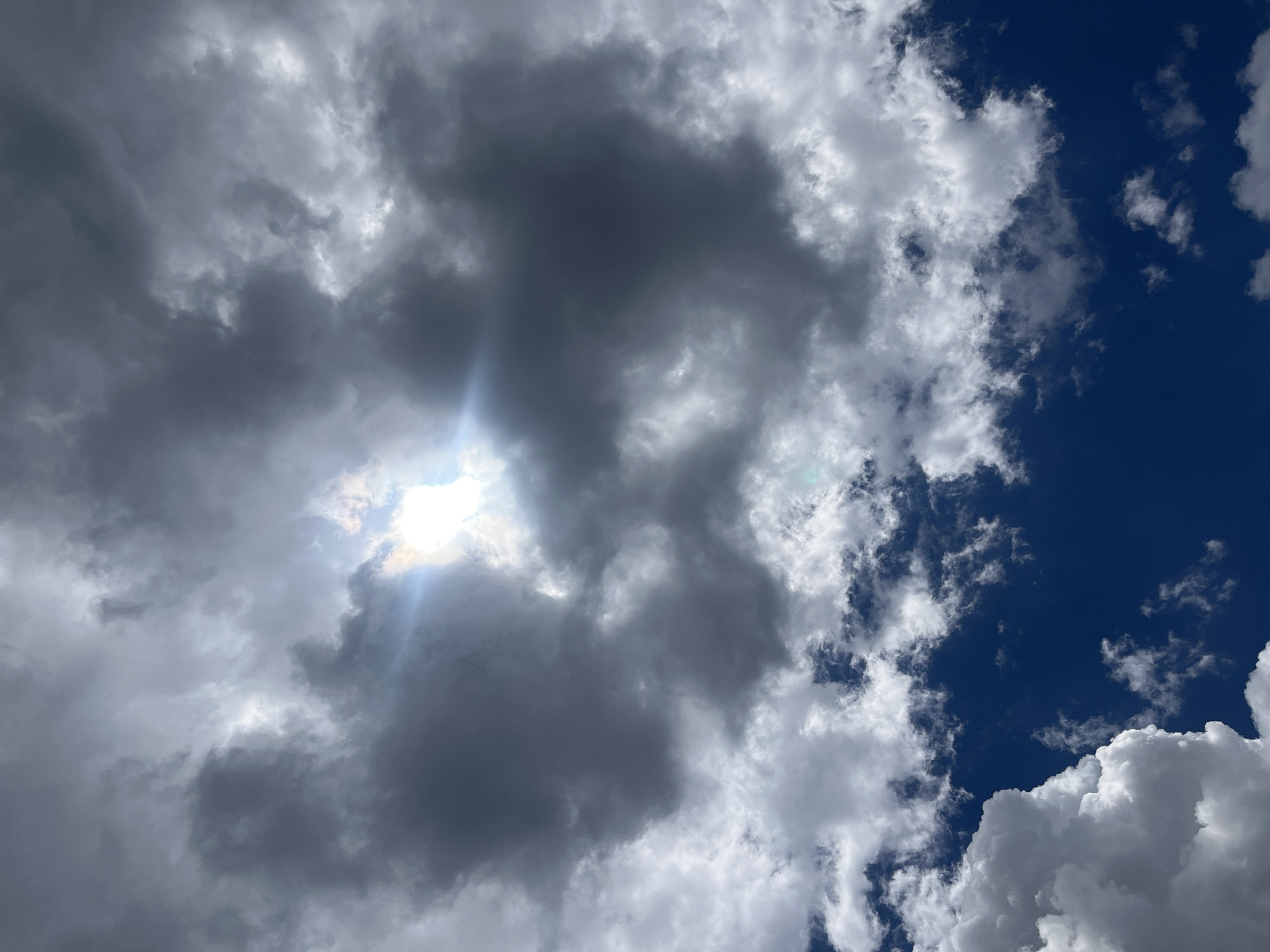 The image depicts a dramatic sky over Jos, Nigeria, characterized by a mix of dark, ominous storm clouds and bright, fluffy cumulus clouds. The sun is partially obscured by a large, dark cloud formation in the upper center of the frame, casting a bright, lens-flared glow through a gap. The intense sunlight creates a stark contrast with the heavy clouds, suggesting an impending storm or the clearing after one. The deep blue of the sky is visible in some areas, particularly towards the bottom of the image where lighter clouds are scattered. The overall impression is one of dynamic weather conditions. There are no people, subjects, or objects other than clouds and sky visible in the image.