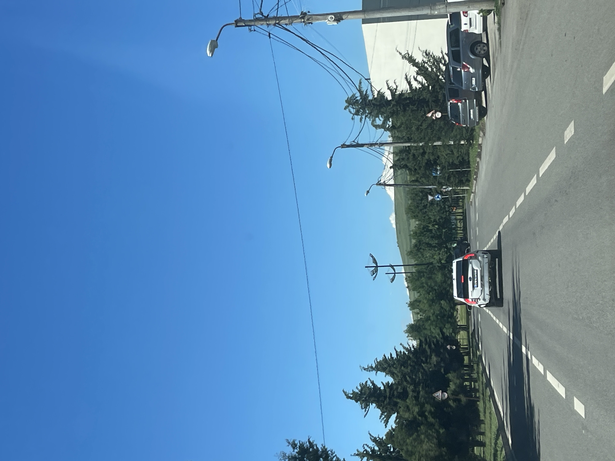 The image shows a street in Tbilisi, Georgia, on a clear sunny day. The perspective is from inside a moving vehicle, looking forward. The road is paved with asphalt and has white dashed lines indicating lanes. There are several cars on the road and parked along the side. Tall green trees line the left side of the road, with a grassy area and more trees in the background. The right side of the road has parked cars and a white building. There are several utility poles with wires running across the scene, and streetlights are visible. In the distance, there are rolling green hills. The sky is a clear, bright blue.