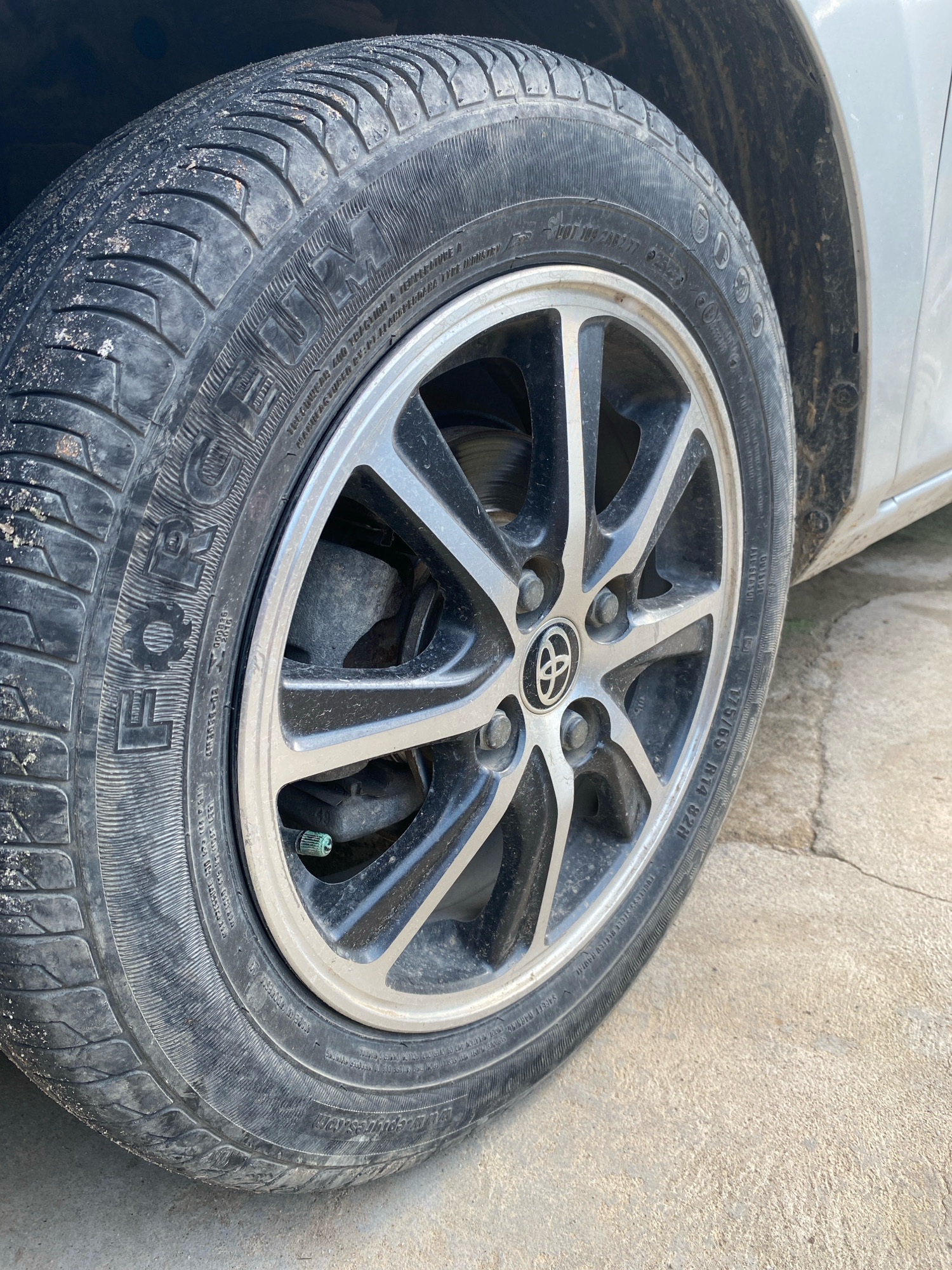The image shows a close-up of a car tire and rim, with the car's body visible in the background. The tire is black and appears to have some dirt or sand on its surface. The rim is silver and black with a multi-spoke design, and features a Toyota logo in the center. The car is parked on a cracked concrete surface, and the area around the wheel well is dirty. The overall scene suggests an outdoor environment, possibly a street or parking lot in Pantai Gading, Indonesia. There are no people or any discernible actions or interactions taking place.