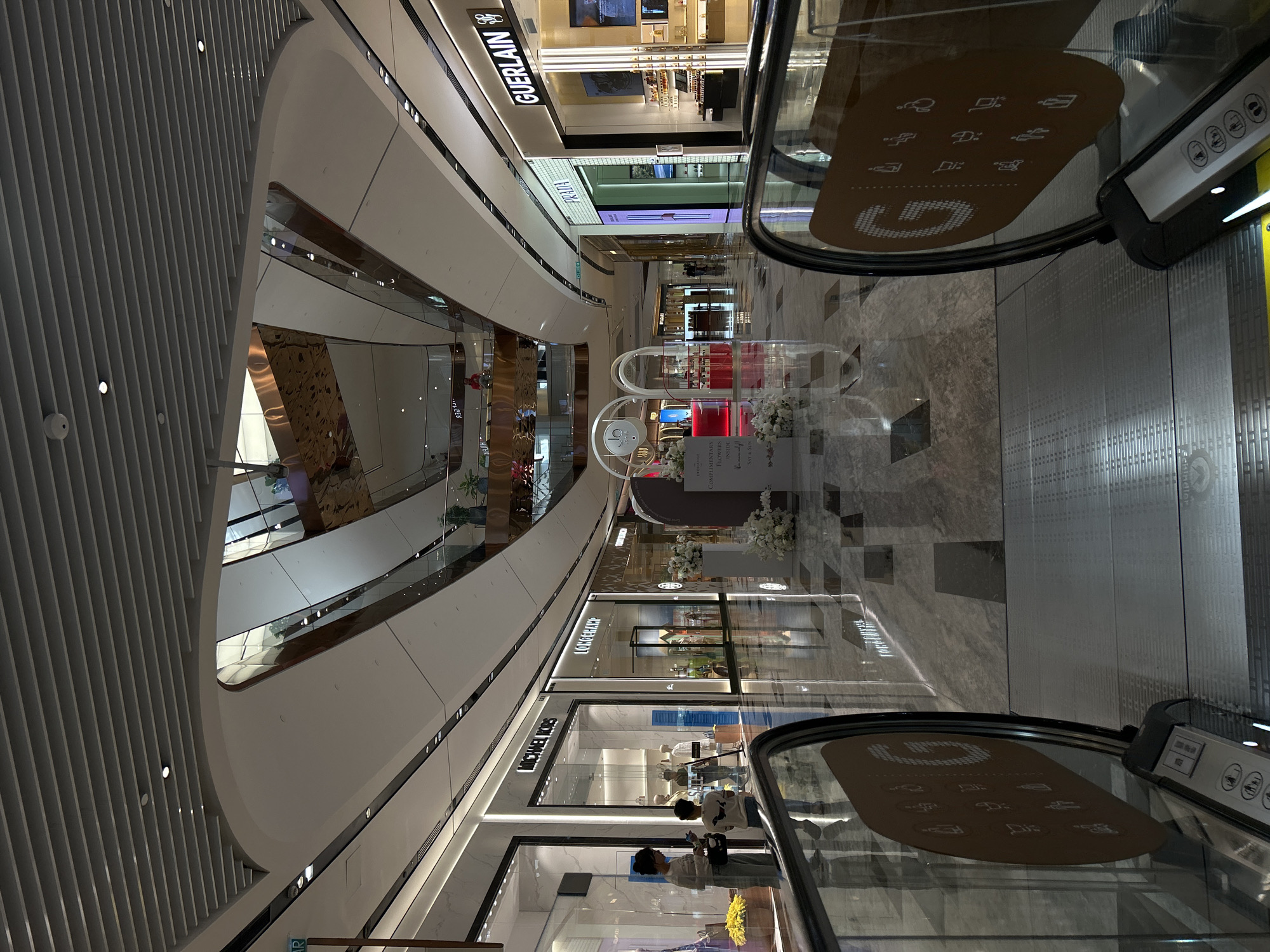The image depicts an interior view of a high-end shopping mall, possibly in Kuala Lumpur, Malaysia, as suggested by the presence of international brand stores such as Guerlain and Michael Kors. The shot is taken from an elevated position, likely on an escalator, looking down at the mall's central atrium. The mall features multiple levels, each with storefronts visible. The architecture is modern with clean lines, white walls, and metallic accents. The flooring appears to be marble or polished stone.
People are visible in the foreground, near the escalators, and in the background, browsing in shops. A display with flowers and what appears to be perfume bottles stands in the mall's central area. Escalators with dark handrails are positioned on either side of the frame, creating vertical lines in the image. The presence of brand stores and the mall's architecture suggests a commercial environment.