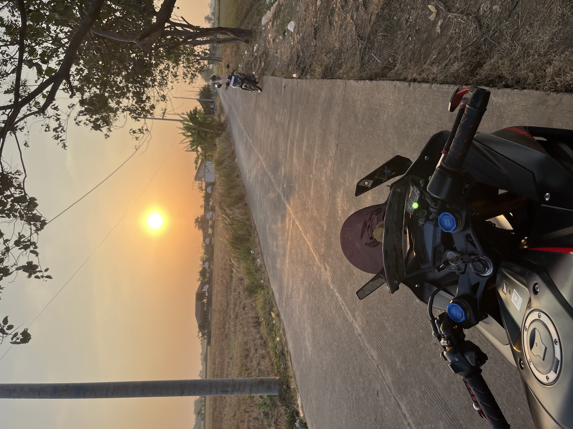 A motorcycle is parked on the side of a paved road in Pecangaan, Indonesia, at sunset. The sun is low in the sky, casting a warm orange glow over the scene. The road is lined with dry grass and trees. In the distance, there are houses and a field. The motorcycle is black and red, with a tinted windshield. The handlebars are visible, with red grips. The seat is black. Another motorcycle is parked further down the road. The sky is clear and the air appears hazy.