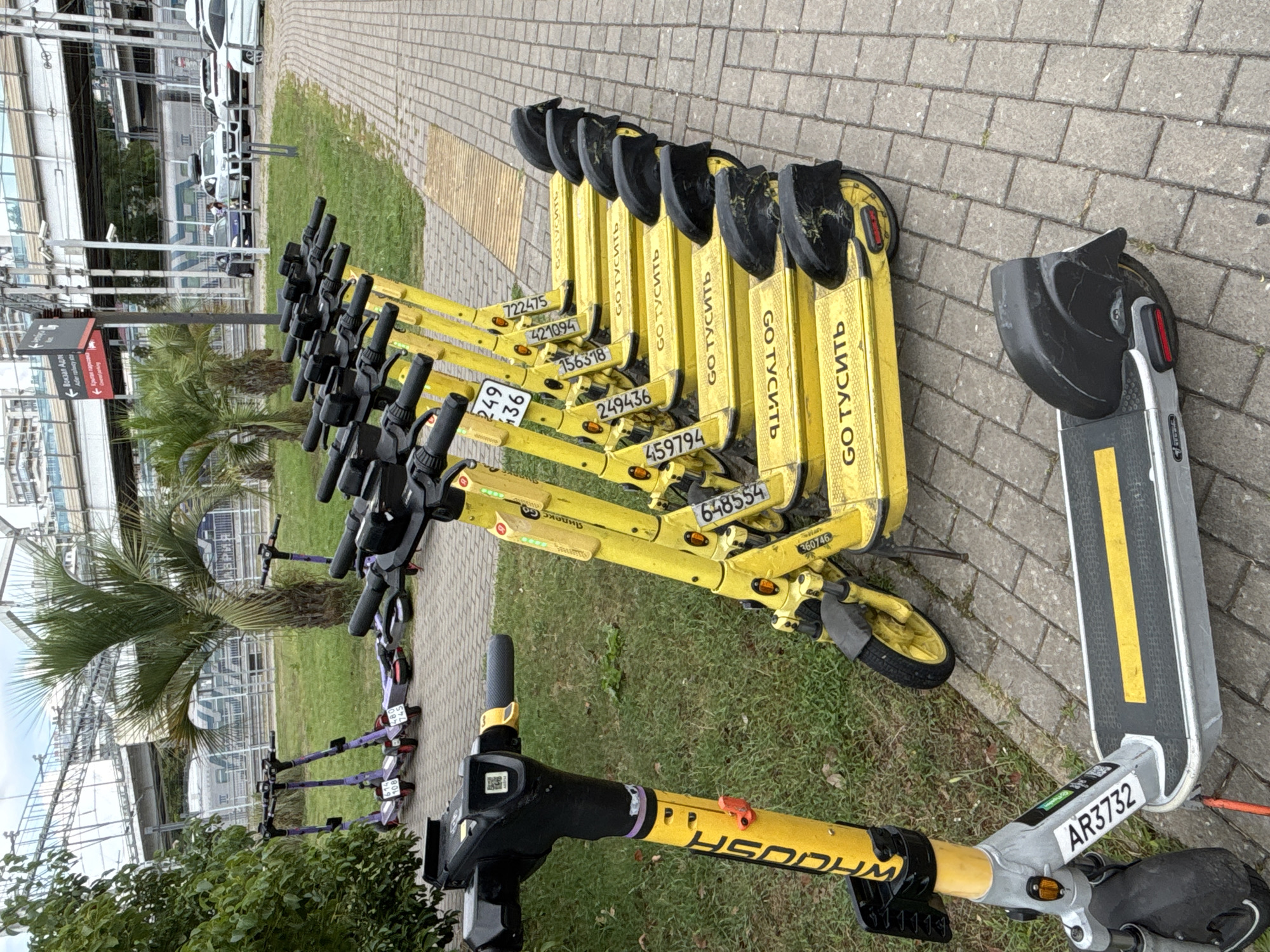 In Sochi, Russia, a row of yellow electric scooters is parked on a brick-paved walkway next to a grassy area. The scooters are lined up neatly, with their handlebars turned to the left. The closest scooter in the foreground is silver and black with a yellow stripe on the deck and a license plate reading "AR3732". The other scooters are predominantly yellow and have markings such as "GO ТУСИТЬ", "Яндекс", and various numbers on their sides. In the background, palm trees are visible, along with a raised railway or bridge structure and a train on tracks. Several cars and people are also present in the background near a signpost with Russian text. The sky is overcast.