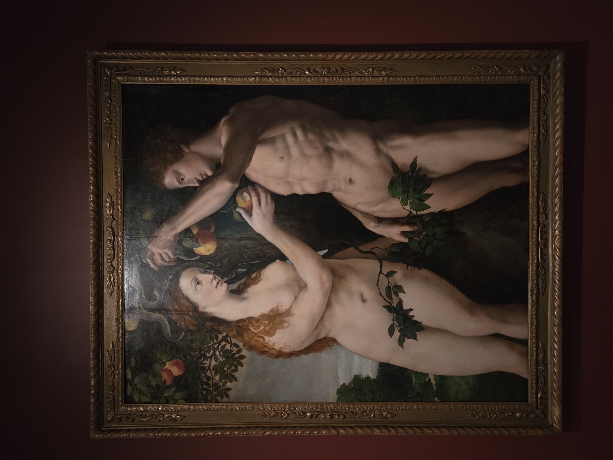 An oil painting in a gilded frame depicts Adam and Eve in a garden. Eve, with long red hair, offers an apple to Adam, who has an athletic physique and curly brown hair. A serpent coils around a branch in the background. Adam holds a branch with leaves partially obscuring his lower body, and Eve has a branch with leaves covering her lower body. The painting is displayed on a dark red wall in Malmö, Sweden, within the Nationalmuseum, which houses a significant collection of art.