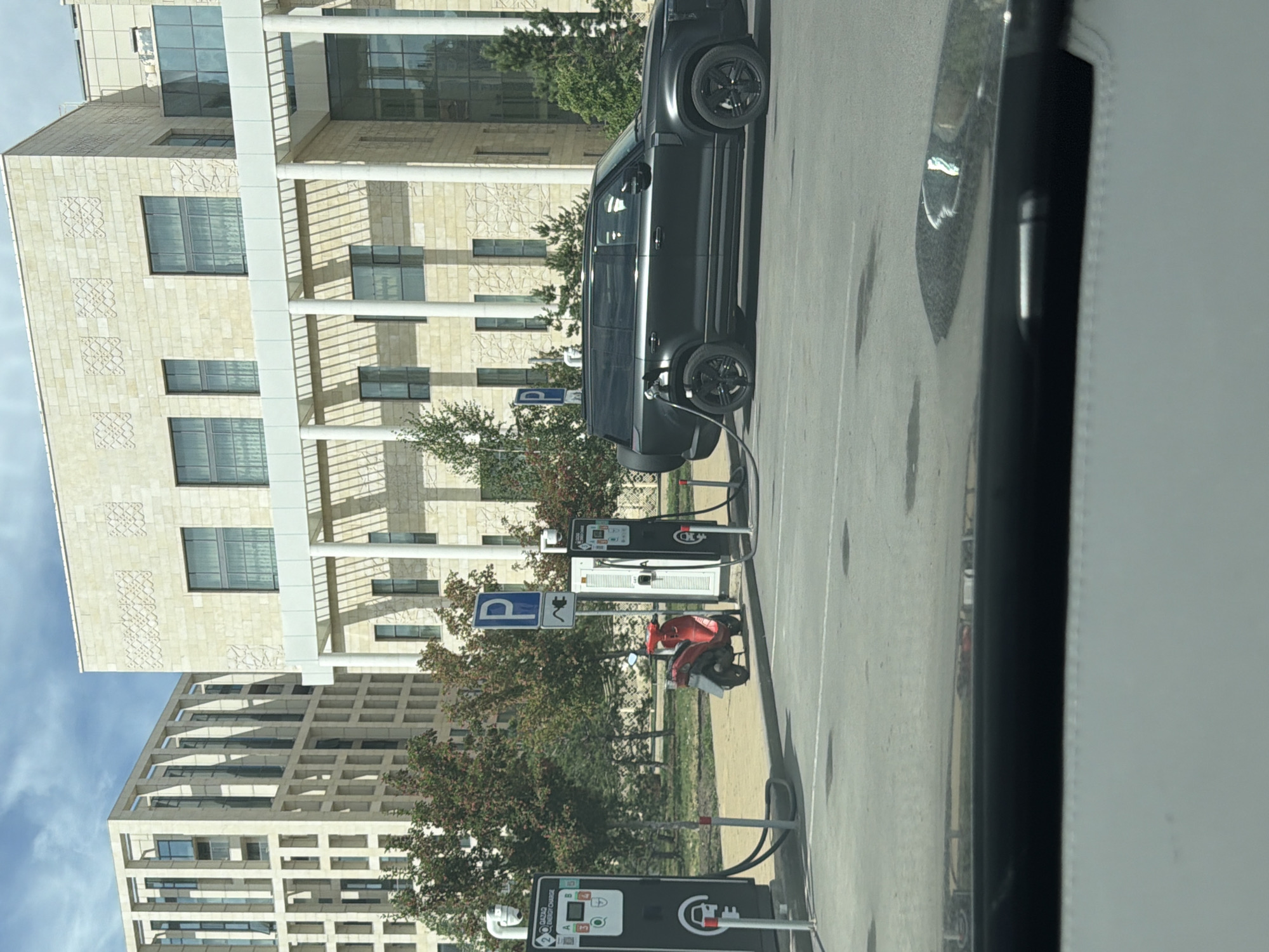 In Astana, Kazakhstan, in front of a modern stone building with patterned facade elements and a white covered walkway supported by columns, an electric vehicle charging station is visible. A dark-colored SUV, likely a Land Rover Defender, is plugged into one of the charging stations. To the left of the SUV, a red scooter is parked next to another charging station. Two charging units are prominently displayed, one with the "GAZAD ENERGY CHARGE" logo. Behind the charging stations, trees and greenery line the area. In the background, a contemporary beige building with a grid-like facade and many windows is visible, adding to the urban architectural context of Astana. The sky above is partly cloudy.