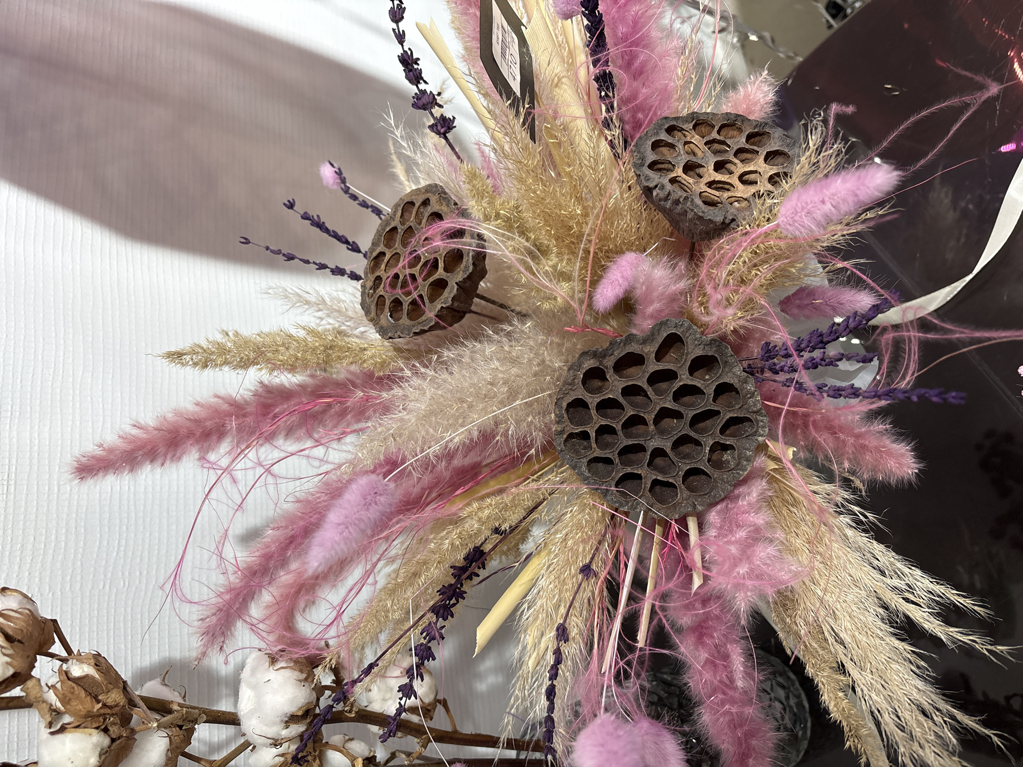 The image shows a dried flower arrangement with pink and beige feathery elements, dark brown lotus seed pods, and sprigs of purple flowers. A branch with cotton bolls is visible in the lower left corner. The arrangement appears to be displayed indoors against a white wall. The specific location in Nizhnevartovsk, Russia, cannot be determined from the image.