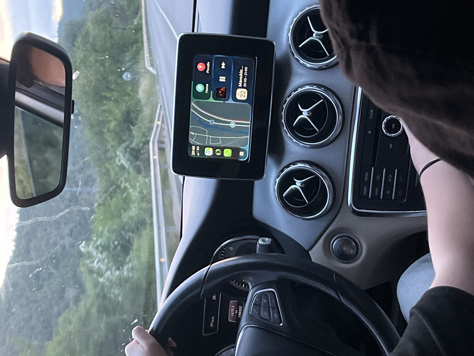 The image shows the interior of a car, specifically the driver's area. A person is holding the steering wheel, and the dashboard is visible, featuring air vents and a display showing navigation and media information. The speedometer reads 77 km/h, and the odometer shows 198502 km. The display also shows "Manuklo 20:00-21:00". Outside the windshield, a road is visible, bordered by green trees and vegetation, suggesting a countryside or suburban setting.