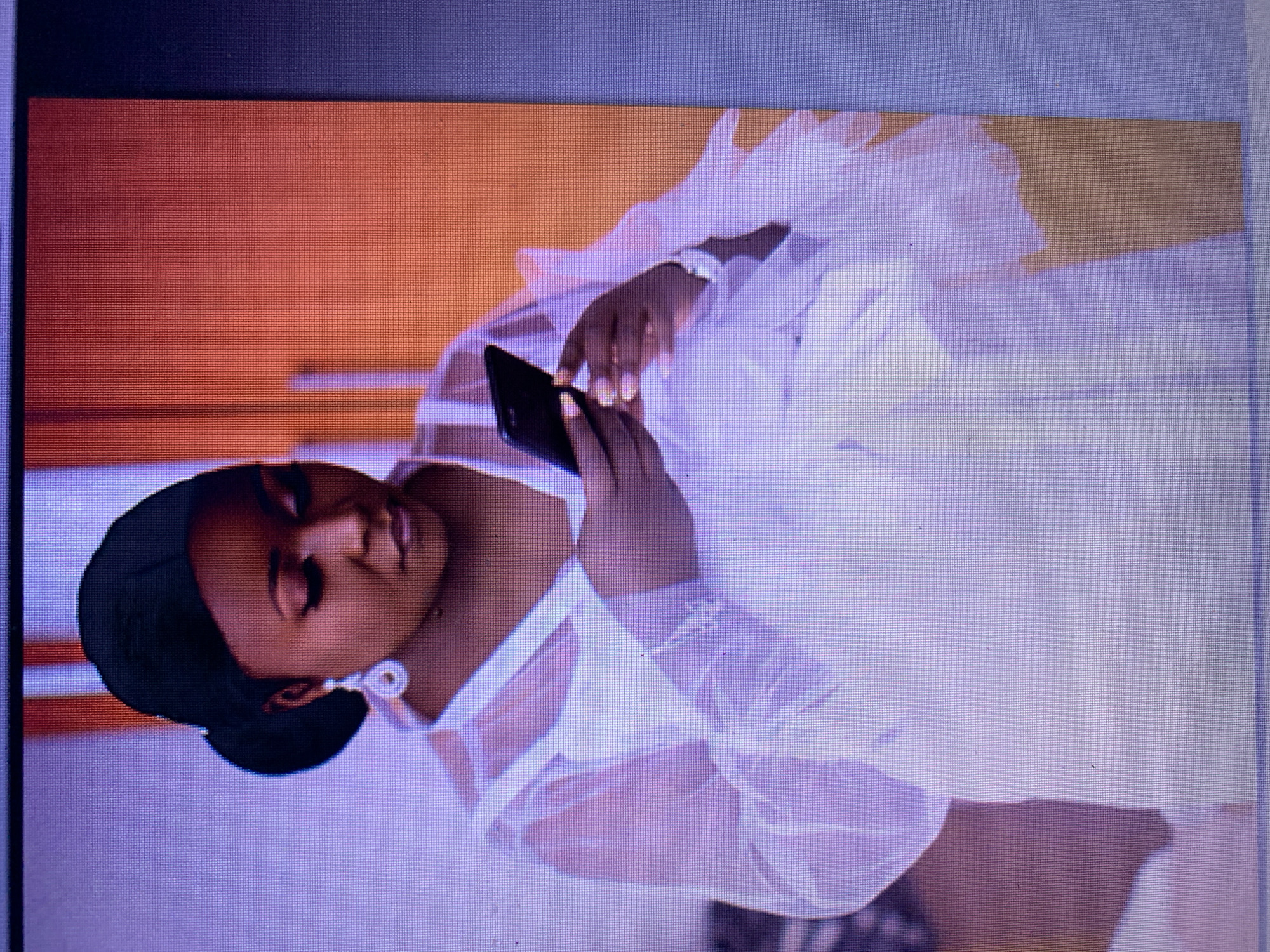A woman is looking down at her phone. She has dark hair pulled back into a bun and is wearing a white sheer robe over a white garment. She has on large teardrop earrings and a watch on her left wrist. The background is out of focus, but appears to be an indoor setting with an orange wall. The image is cropped and appears to be taken from a screen.