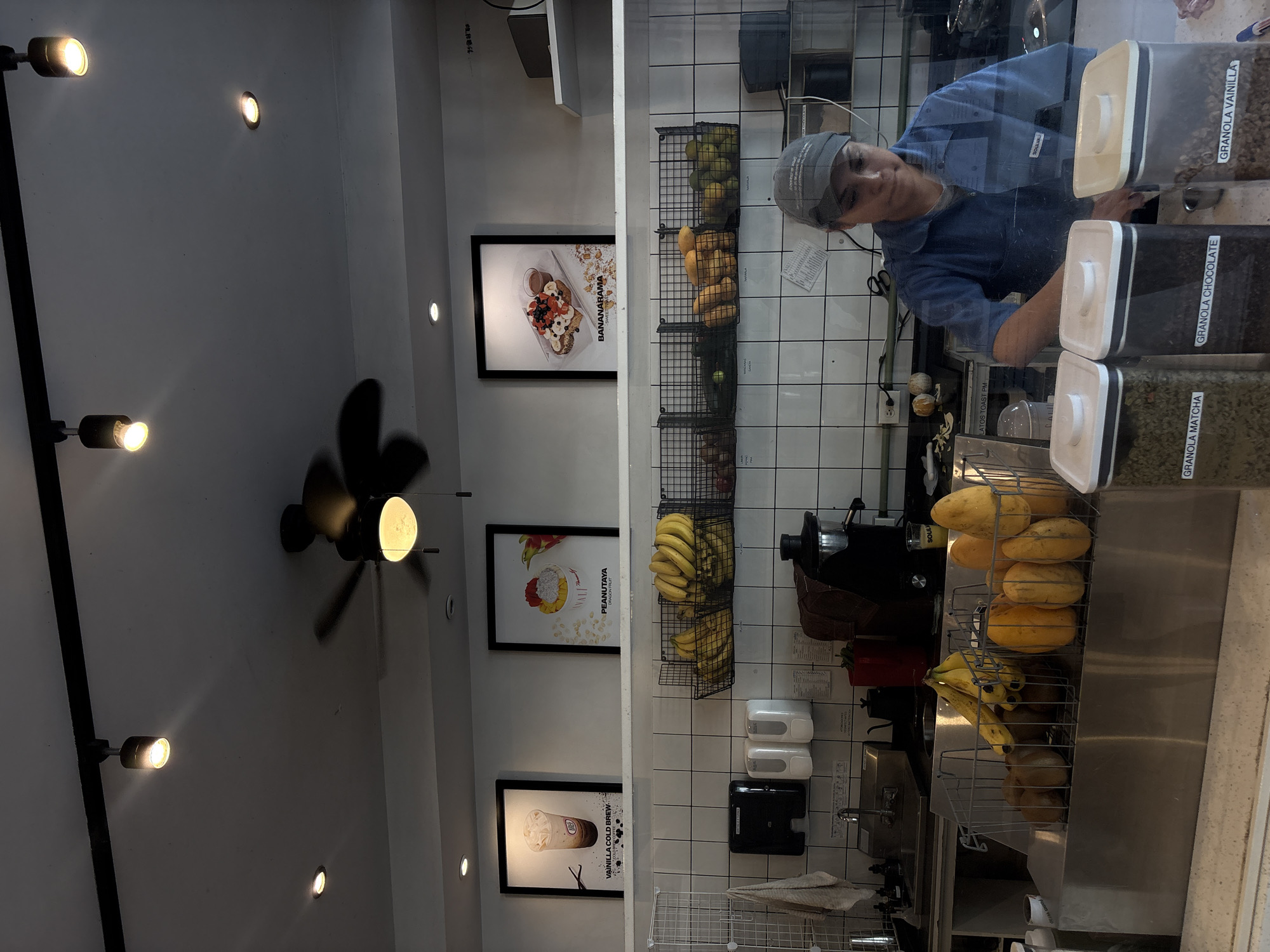 The image captures an indoor scene, likely a cafe or smoothie bar, with a worker behind a counter. The main subject is a person, appearing to be a woman, wearing a blue shirt/uniform and a gray cap or hairnet, looking towards the viewer with a neutral expression. She is positioned behind a glass partition, with her reflection slightly visible.
The counter area is equipped for preparing fresh food or drinks. Several types of fruit are visible in black wire baskets, including bunches of ripe bananas, papayas, and what appear to be oranges or limes. On the countertop to the right, there are clear plastic containers with white and black lids, labeled in Spanish for different granola flavors: "GRANOLA VAINILLA" (Vanilla Granola), "GRANOLA CHOCOLATE" (Chocolate Granola), and "GRANOLA MATCHA" (Matcha Granola). Kitchen equipment, such as blenders or juice presses, and a sink area are also present. The wall behind the counter is covered in white tiles.
The left side of the scene shows a white wall adorned with framed posters advertising menu items. One poster features a dish called "BANANARAMA," another "PEANUTAYA DRAGON FRU" (likely Dragon Fruit), and a third "VAINILLA COLD BREW." The lighting consists of multiple artificial sources: track lighting fixtures, recessed ceiling lights, and a ceiling fan with an integrated light. Some of these lights cast bright glows on the otherwise neutral-toned walls. The overall impression is that of a clean, functional commercial space.
The visible text, including "BANANARAMA," "VAINILLA COLD BREW," "PEANUTAYA DRAGON FRU," "GRANOLA MATCHA," "GRANOLA CHOCOLATE," and "GRANOLA VAINILLA," along with "SOUL" and "SOULKK" on equipment, suggests a food and beverage establishment. The Spanish language on the labels further supports the context of Puebla, Mexico.
