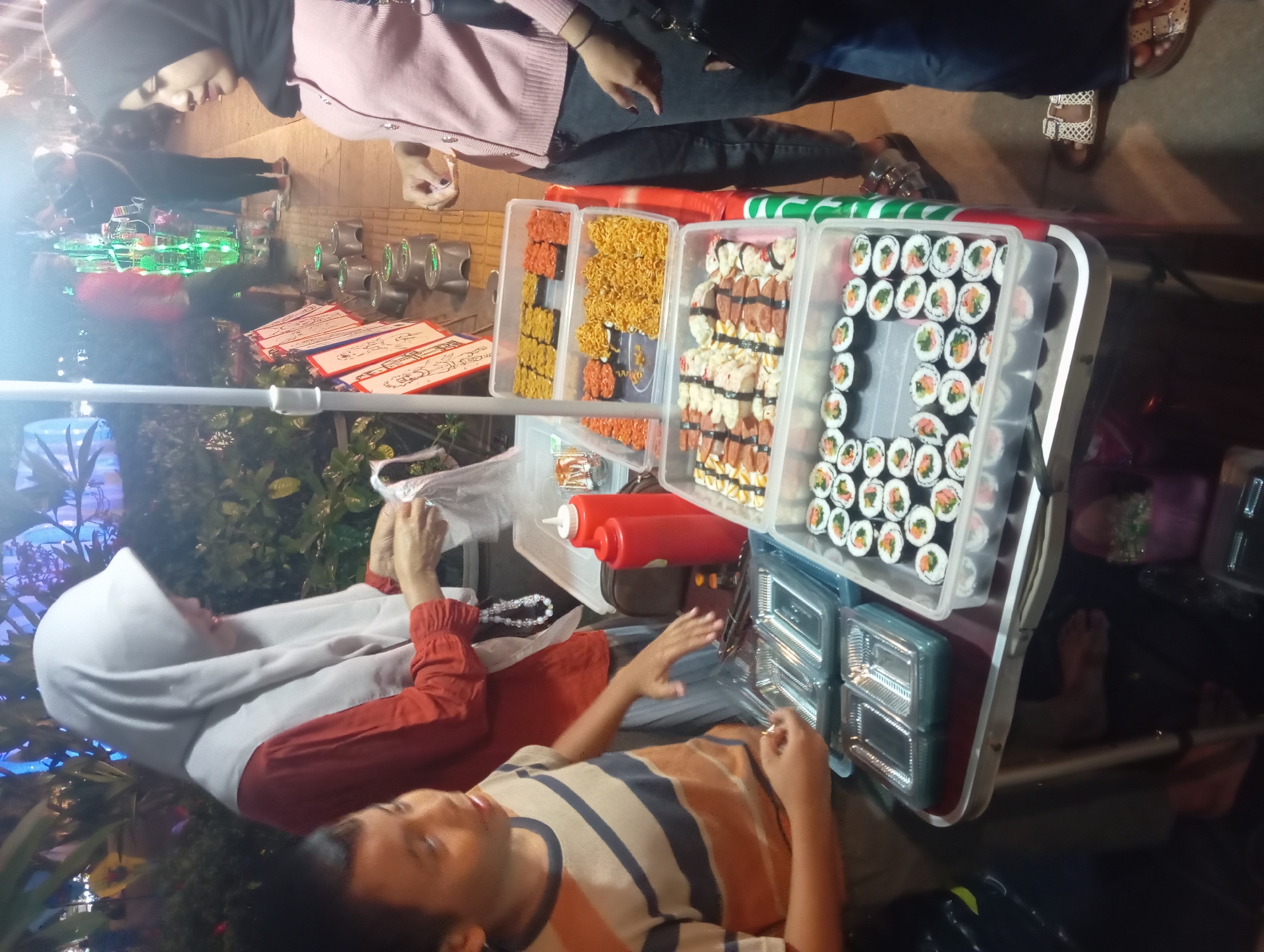A street vendor in Purwokerto, Indonesia, is selling sushi rolls and other food items from a portable stall. The vendor, a young woman wearing a hijab and a pink cardigan, is interacting with a child who is reaching towards the food. The stall is set up on a sidewalk at night, with lights illuminating the scene. In the background, other people are visible, as well as various food displays and decorations. The food items being sold include sushi rolls with various toppings, as well as what appear to be fried noodles and other savory snacks. The scene depicts a lively night market atmosphere in Purwokerto.
