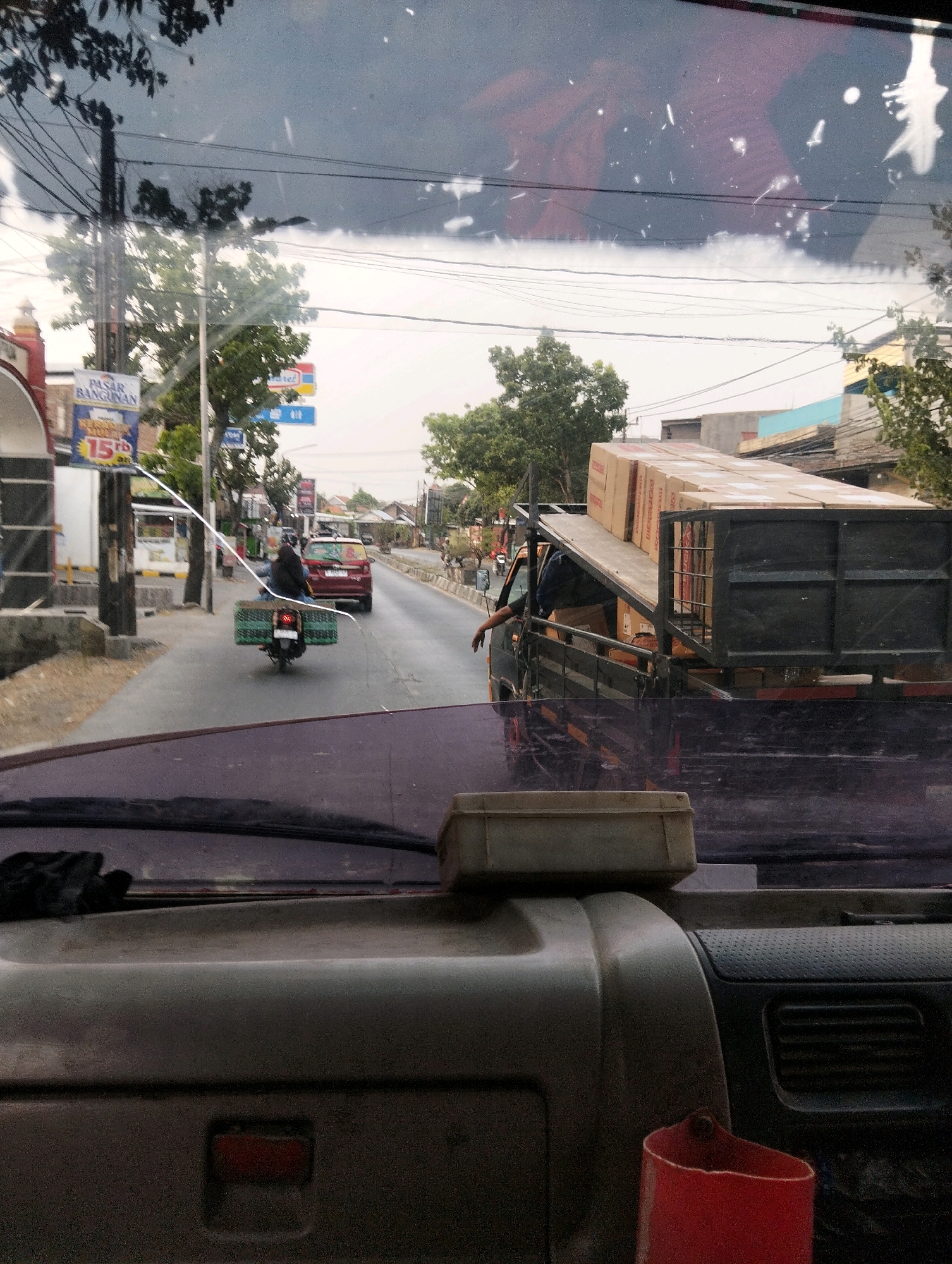 The image captures a street scene in Semarang, Indonesia, viewed from the interior of a vehicle. The foreground shows the dashboard and windshield of the vehicle, with a red container and a grey plastic box visible. Outside, a road stretches into the distance, flanked by buildings and trees. On the road, a red car is visible, as is a motorcycle carrying a cargo of what appears to be cardboard boxes. A flatbed truck, also laden with boxes, is positioned on the right side of the road, with a person's arm resting on its side. Billboards, including one that reads "PASAR BANGUNAN" (Building Market), are present along the street, indicating commercial activity. Overhead, power lines crisscross the sky, suggesting an urban or semi-urban setting. The sky is overcast.