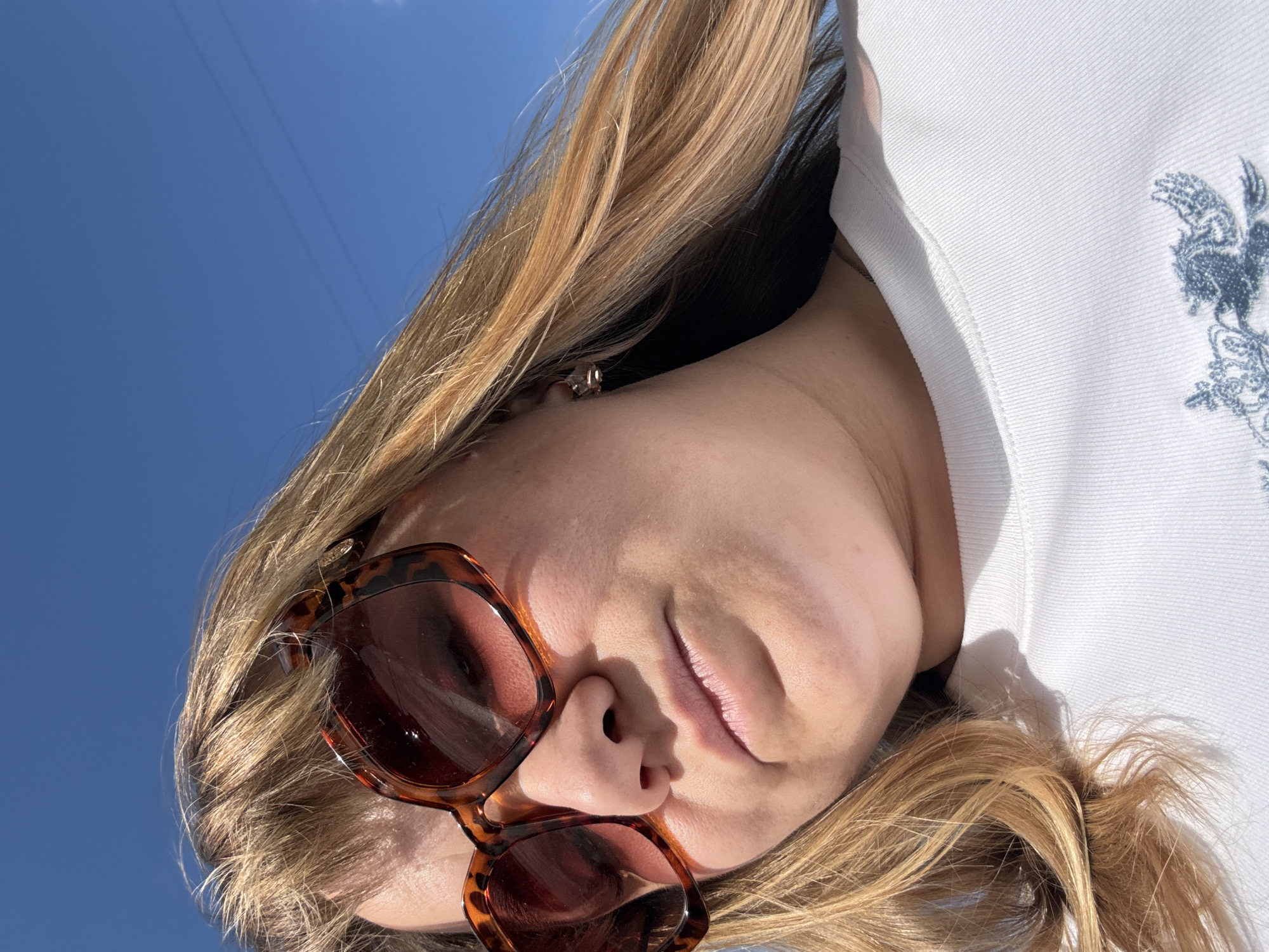 This media file is a close-up, low-angle selfie of a young woman with blonde hair. She is wearing large, tortoiseshell-patterned sunglasses and a white hoodie with a blue embroidered design on the chest. The background is a clear, bright blue sky with two thin, dark lines, possibly power lines, stretching across it. The lighting suggests it is daytime, likely a sunny day, casting shadows on the woman's face and neck. Her hair is flowing around her face and neck, and the sunlight highlights the golden tones in her hair. She appears to be outdoors, possibly in Stary Oskol, Russia, given the location context. Her facial expression is relaxed, with her lips slightly parted and a gentle smile. The overall mood is casual and summery.