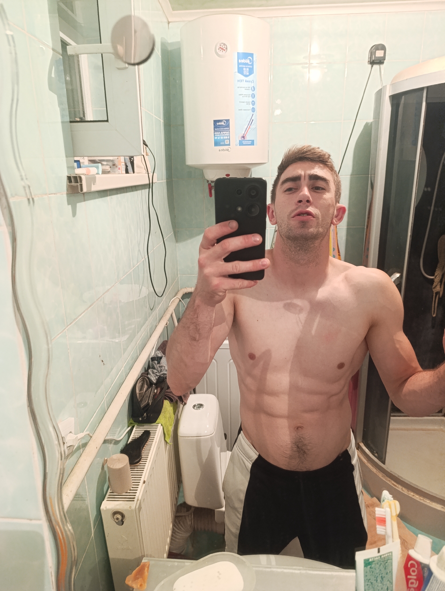 The image is a mirror selfie taken in a bathroom. A shirtless man with visible abs is posing for the camera, holding a smartphone. The bathroom appears to be in a residential setting, with tiled walls, a water heater, a radiator, a toilet, and a shower enclosure visible. There are also toiletries and other bathroom items on shelves and surfaces. The man is wearing black and white shorts. The location is specified as Fastiv, Ukraine.