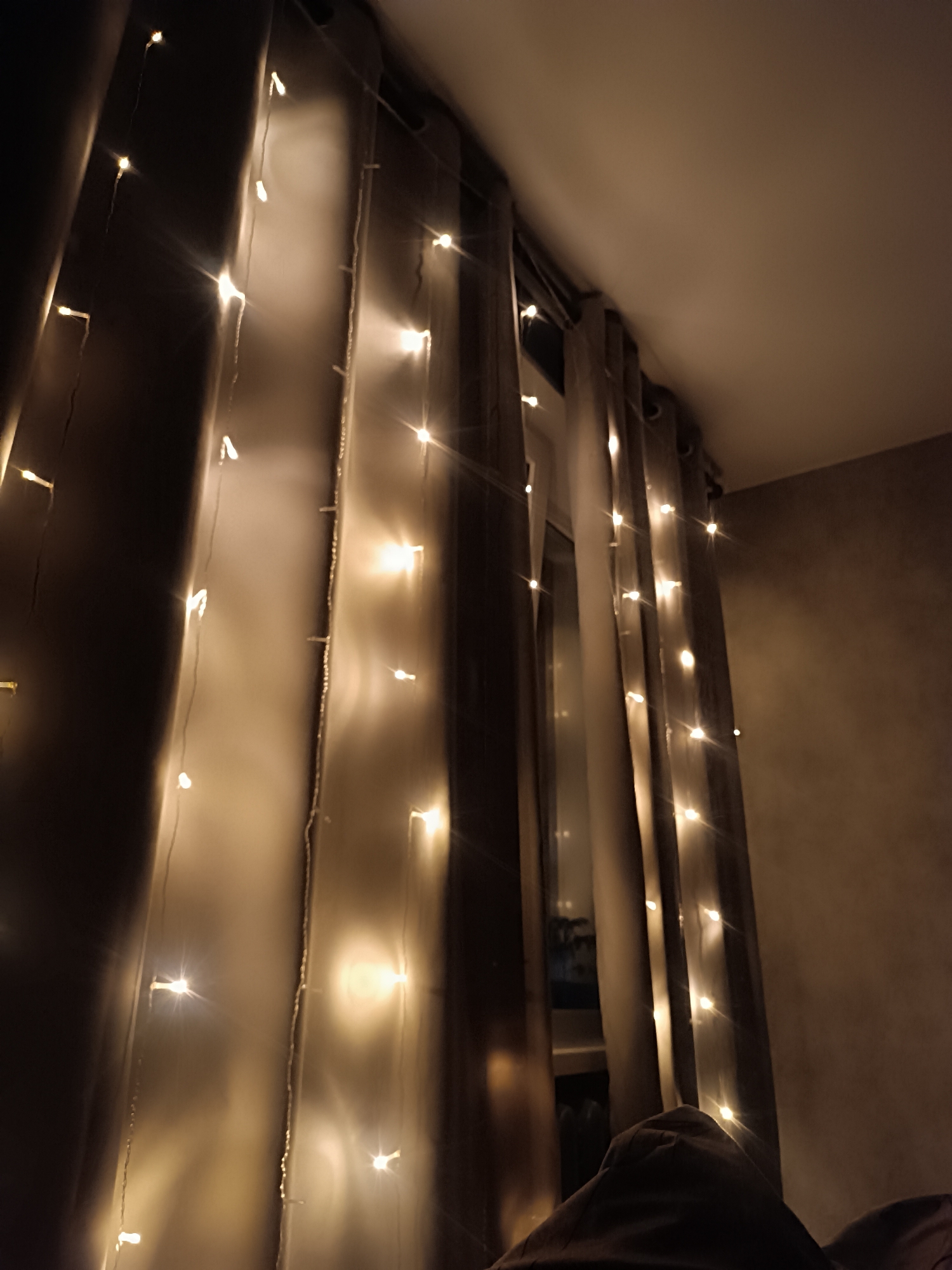 A low-angle, indoor shot depicts a darkened room illuminated by a string of fairy lights hung on curtains. The curtains are draped along a window frame, creating a soft, glowing effect from the light bulbs. The wall is dark, with a sheen. The ceiling is white. The photo is taken from below, so the top of the bed is visible.