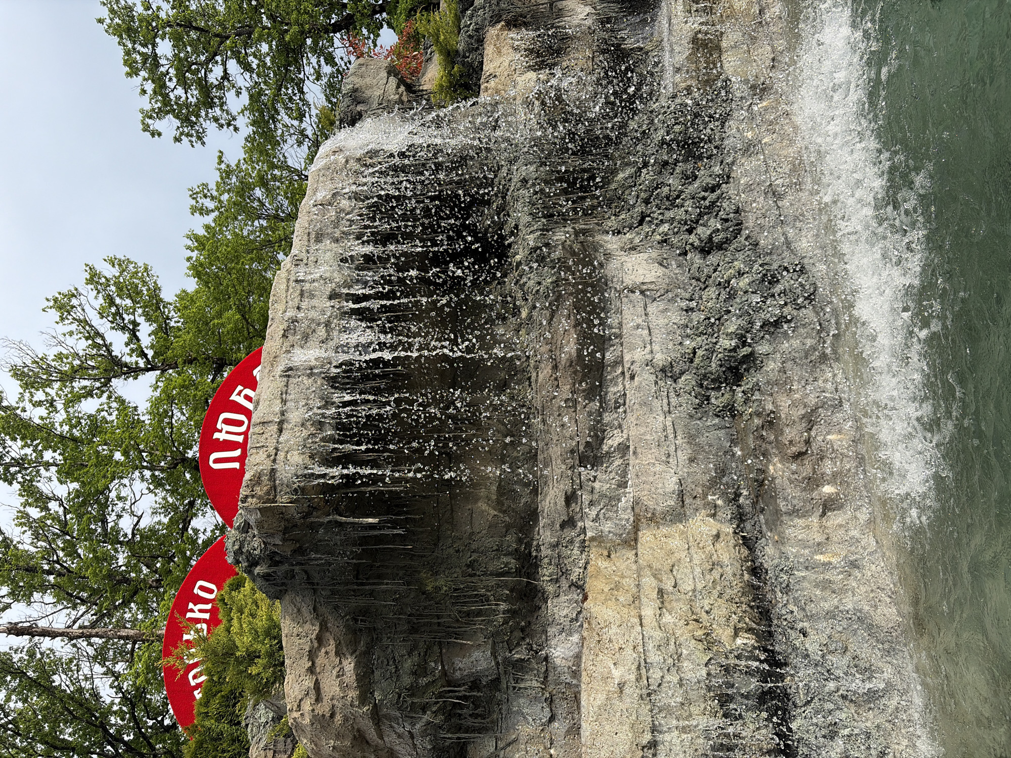 The image shows a waterfall cascading over rocks. Vegetation, including trees, surrounds the area. A red sign with white Cyrillic lettering is partially visible behind the waterfall and the rocks. The sign, with the letters "Любовь" and "Ключко", indicates the location is in the city of Goryachiy Klyuch, Russia.