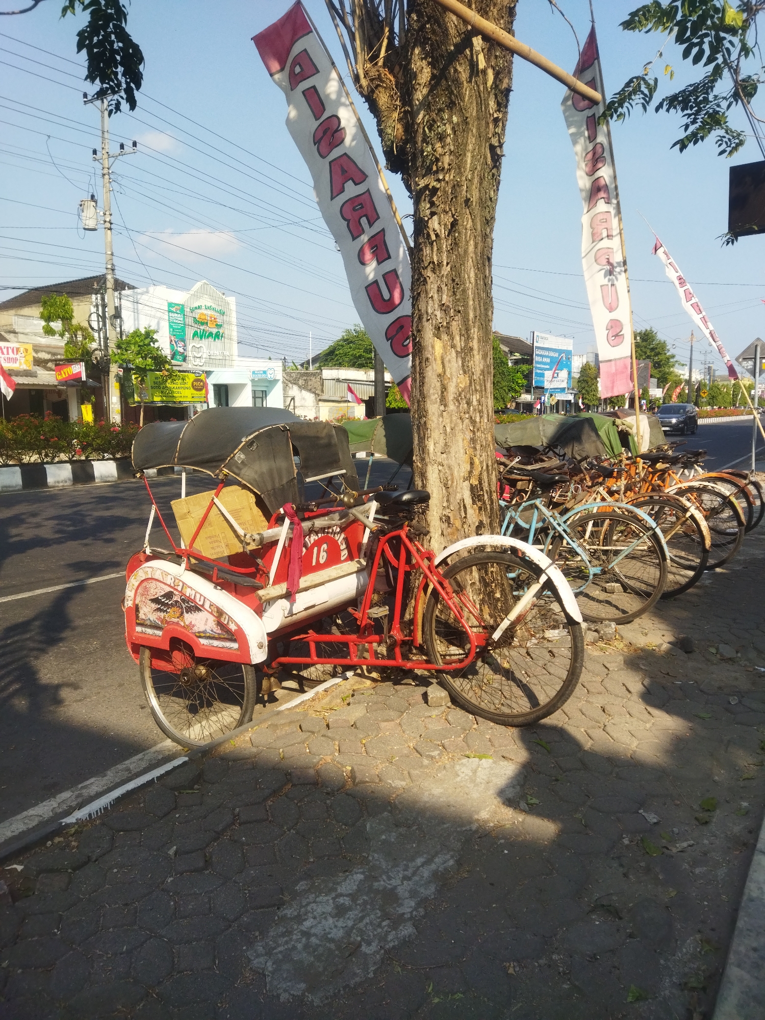 This is a medium shot, eye-level, outdoor photograph captured in Sukoharjo, Indonesia, on a clear day with blue sky. The scene depicts a street with several parked bicycles and a pedicab. In the foreground, a red and white pedicab with a black canopy is prominently displayed. It has decorative painted elements on its side, including an eagle motif. A cardboard box is visible inside the pedicab. A bicycle with a white front fender is parked to the left of the pedicab, facing away from the viewer. Behind the pedicab, a row of several bicycles is parked along a sidewalk. These bicycles are of various colors including blue, yellow, and orange, and appear to be old and weathered. A large tree with textured bark stands behind the pedicab and the row of bicycles, with its branches extending towards the top of the frame.
Several long, white banners with red lettering are hung vertically from poles or wires, draped around the tree and extending upwards into the sky. The lettering on the banners is in Indonesian and appears to be advertising or decorative. In the background, there are buildings, power lines, and utility poles, indicating an urban environment. The buildings include a light-colored structure with a green and white facade and signage. A black car is visible driving on the road in the distance. The ground is paved with hexagonal cobblestones in some areas and asphalt in others, with shadows cast by the objects in the scene due to the bright sunlight. The overall impression is a quiet street scene in a town or city.