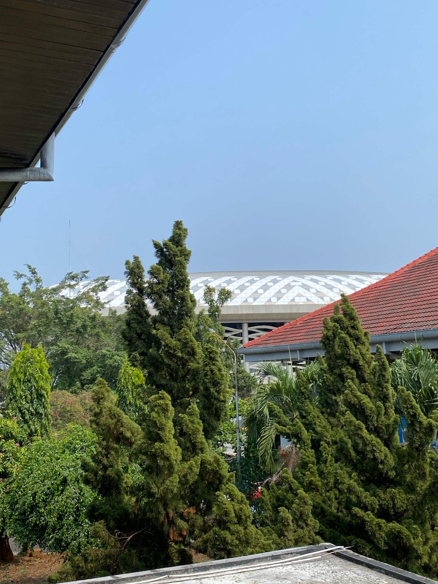 The image captures a daytime view of trees and buildings under a clear blue sky. In the foreground, a portion of a building's roof is visible, along with a gutter system. Beyond this, a dense arrangement of various trees, primarily evergreen conifers and leafy deciduous trees, fills the middle ground. Behind the trees, a modern building with a distinctive white, patterned roof and a red-tiled roof are prominent. The white roof appears to be part of a large structure, possibly a stadium or convention center, with a geometric pattern of alternating light and dark diamonds. The red-tiled roof is attached to a lower building. The overall scene suggests an urban or suburban environment in Semarang, Indonesia, likely with a public facility or event space visible in the background. No people are discernible in the image.