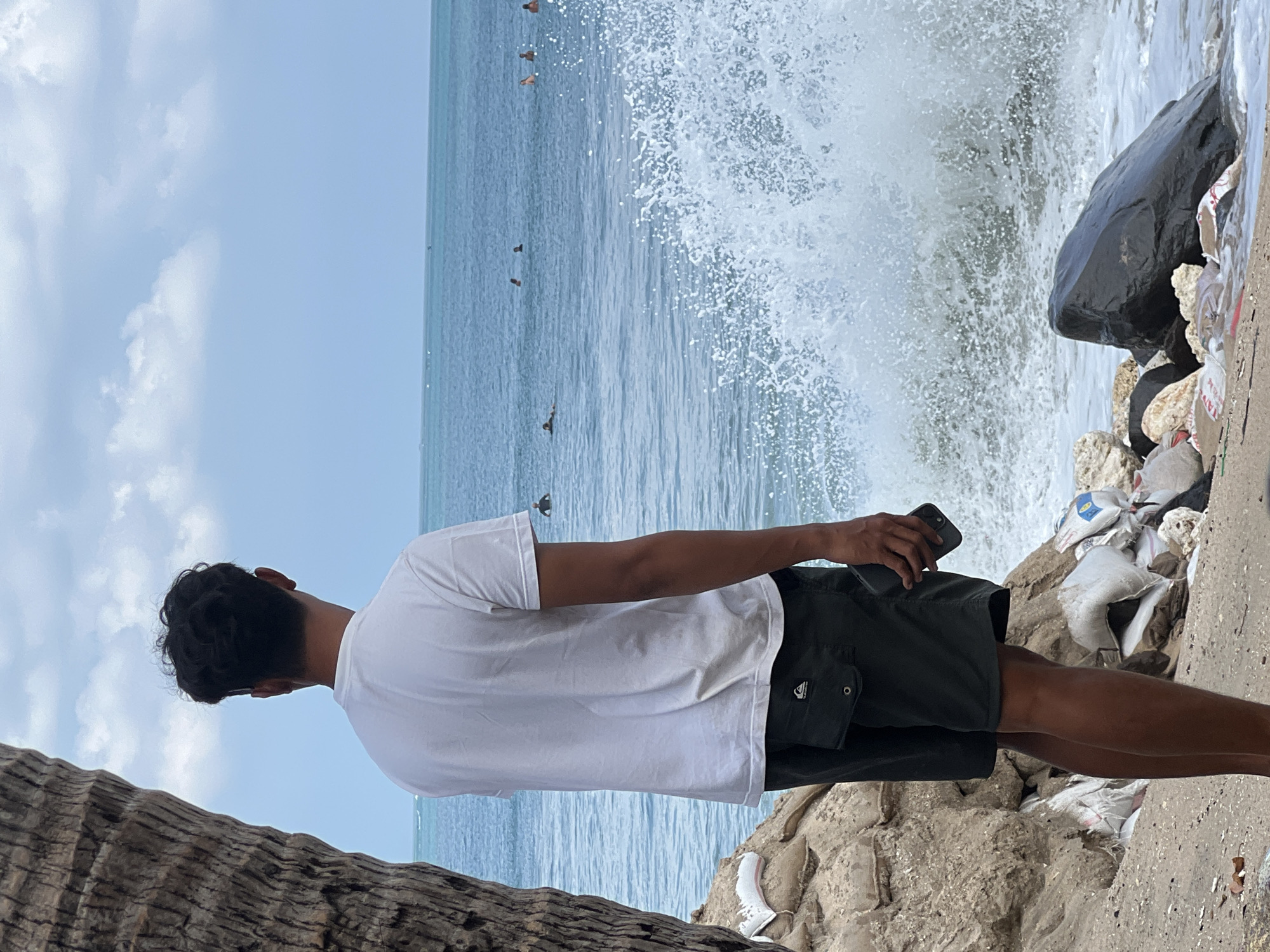 A man stands on the beach in Kuta, Indonesia, facing the ocean. He is wearing a white t-shirt and dark shorts, and holding a smartphone in his right hand. The man is positioned to the left of the frame, with his back mostly to the camera. To his left, a large tree trunk dominates the foreground. The beach is sandy and strewn with various debris, including plastic bags and rocks. The ocean is blue with white waves crashing against the shore. In the distance, several people can be seen swimming or surfing. The sky is partly cloudy.