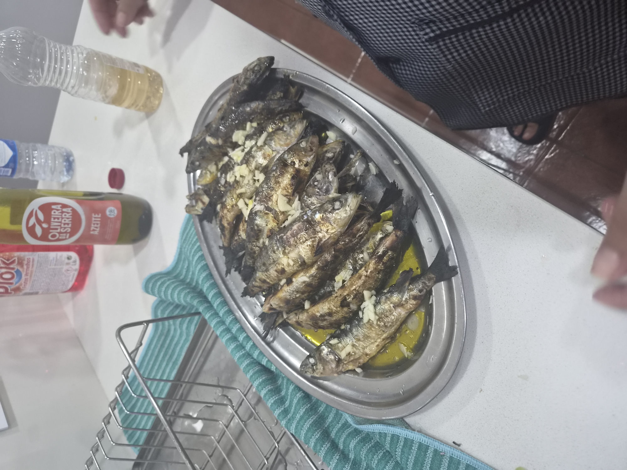 The image captures a close-up, eye-level view of a metallic platter filled with grilled sardines, arranged in rows and generously topped with chopped garlic and olive oil. The sardines are whole, with their skin charred in places, and their tails are pointed upwards in some instances. The platter itself sits on a speckled white countertop, next to a metallic dish rack holding a green and white striped towel.
In the background, on the same countertop, are several items: a red bottle with a white label that reads "Piok," a green glass bottle with a label indicating "OLIVEIRA DA SERRA AZEITE" and "AZEITE" in bold text, and two clear plastic bottles, one containing a light-colored liquid, and another with a blue cap. A small red cap is also visible on the countertop.
The scene appears to be an indoor kitchen or dining area. The floor in the foreground is a reddish-brown tiled surface. A person's legs are visible on the right side of the frame, wearing checkered pants and dark sandals, suggesting they are seated or standing near the table. The overall impression is a prepared meal, likely served in a casual setting. While specific details about Barcelos are not directly evident from the image, the presence of Portuguese branding on the olive oil bottle suggests a connection to Portugal, where Barcelos is located.