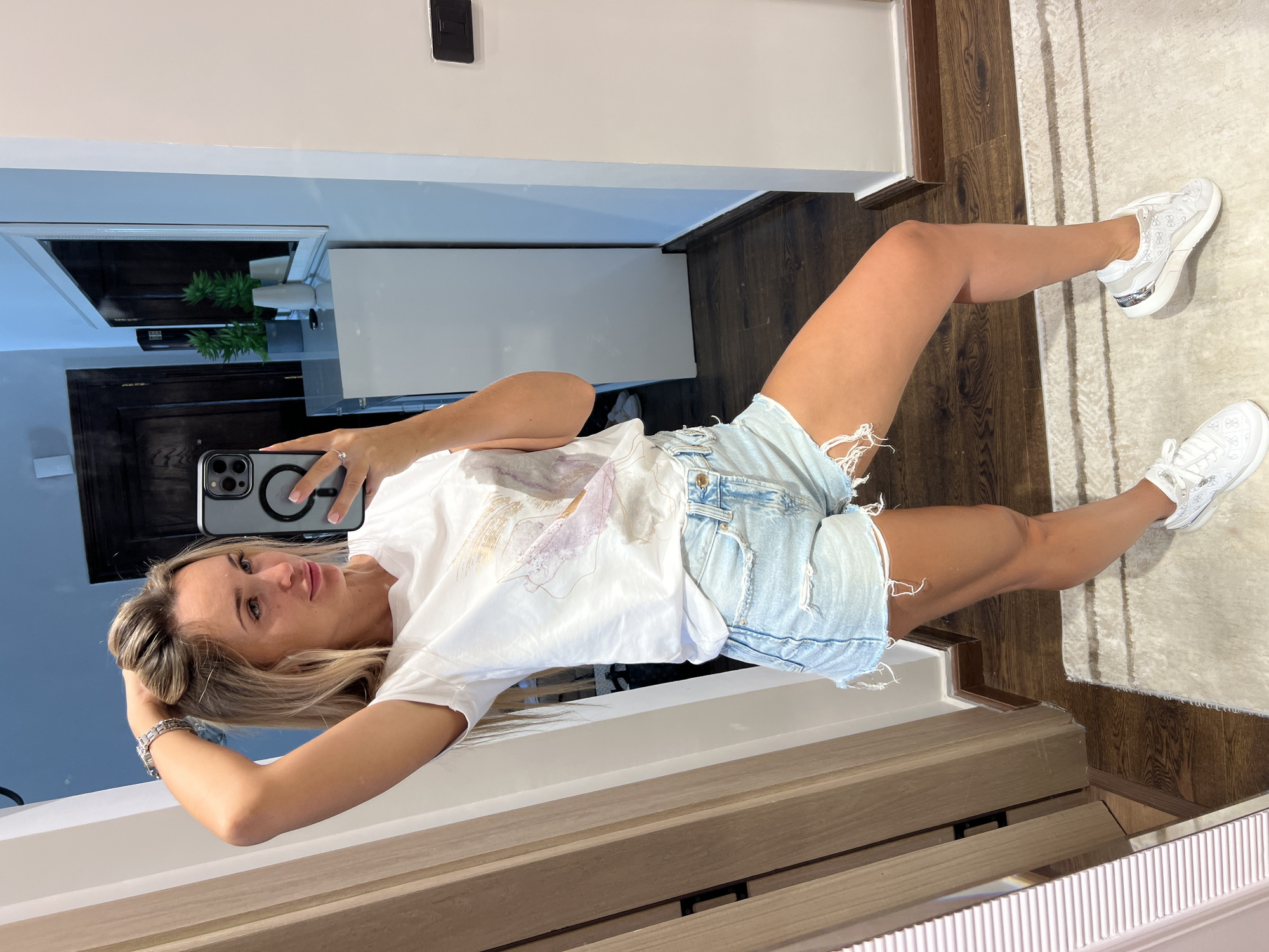 The image shows a woman taking a mirror selfie in what appears to be a well-lit interior space. She has blonde hair, is wearing a white t-shirt with a graphic print, light-wash denim shorts, and white sneakers. Her left arm is raised and her hand is in her hair. She is looking at her reflection in a phone screen. The background shows a doorway, a mirror reflecting a dresser with plants, and a carpeted floor. There are no visible elements that specifically identify the location as Sharm El Sheikh, Egypt.