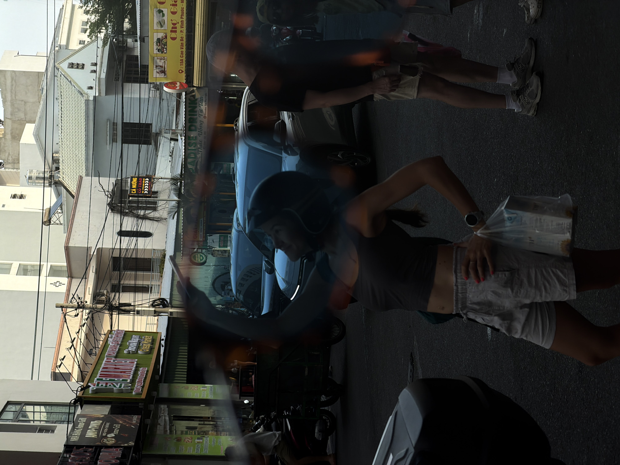 The image depicts a street scene, likely taken from inside a vehicle due to the blurry foreground suggesting movement or being behind glass. A woman wearing a helmet, a gray top, and shorts, is seen holding a plastic bag and walking in the middle of the frame. Another person, a male, also walks in the background, partially obscured. Cars are parked along the side of the road. Signage and buildings are visible in the background. One sign reads "NHÀ HÀNG Amur-Gill" and "АМУР-ГРИЛЬ", suggesting a Russian restaurant. Another sign indicates "Cresp Market" and "Paradise Drink." "Chợ Giả" is written on another sign. The location is indicated on the sign as 15A Cao Van Bé - P. Vinh Phuoc.