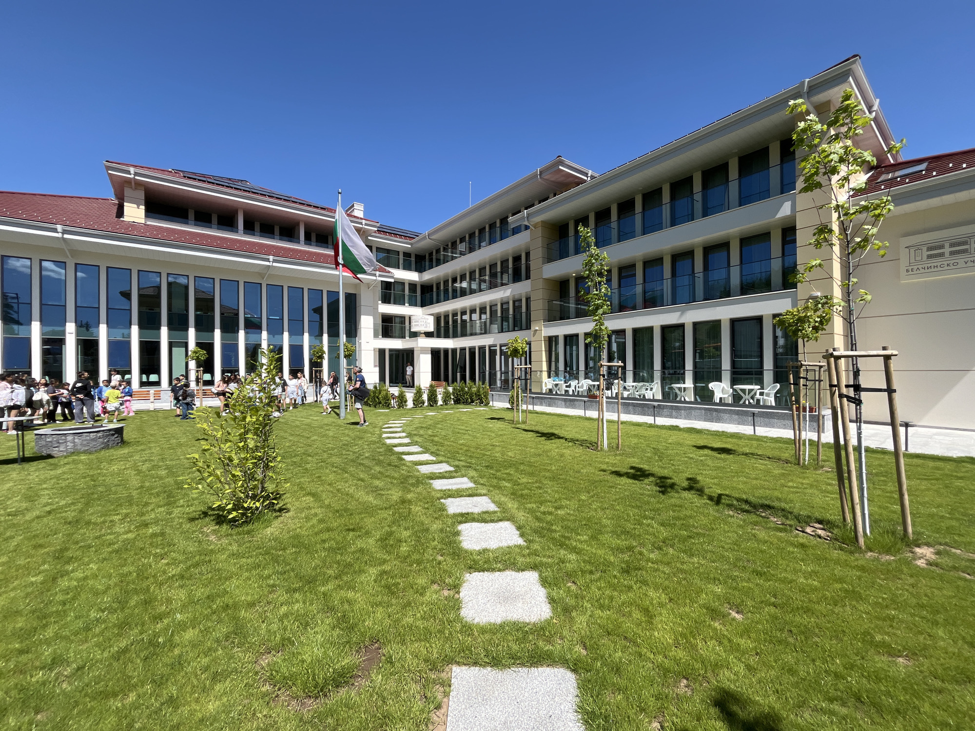 The image depicts the grounds of the Belchinski Uchiha school in Belchin, Bulgaria. The architecture is modern, with a mix of glass and white-painted facades. A lawn stretches in the foreground, intersected by a path of square stone pavers. In the background, the Bulgarian flag stands tall. A small group of people gathers near the building on the left, while outdoor tables and chairs suggest a dining or relaxation area. Young trees are planted in the grass, supported by stakes.