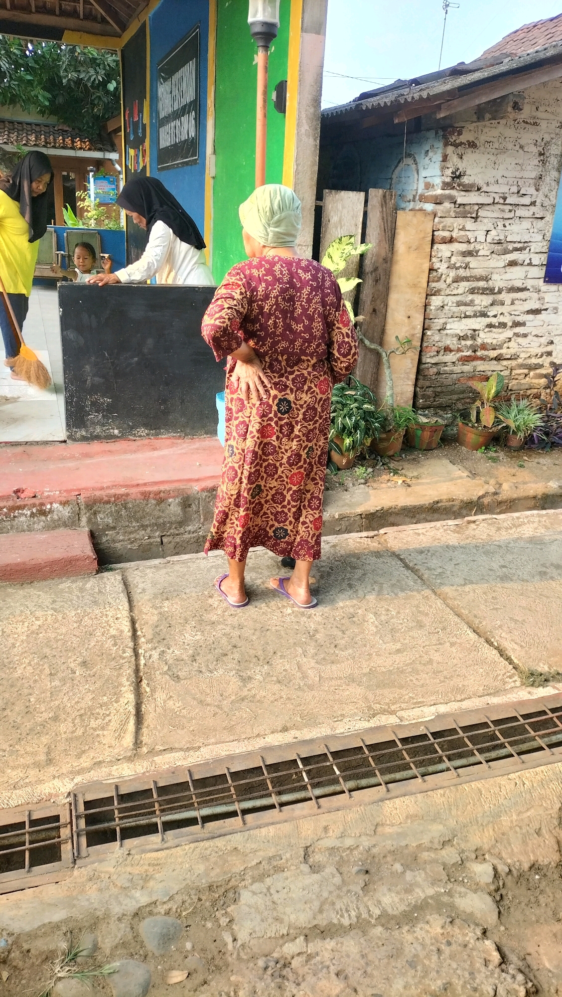 The image depicts a street scene in Subah, Indonesia, under bright daylight. In the foreground, a person wearing a patterned maroon dress and a light green headscarf stands with their back to the camera, hands on their hips. The individual is wearing purple flip-flops. To the left, a grate runs along the edge of the sidewalk. In the background, there is a small building with blue and green painted walls. Inside the building, a person in a white top and black headscarf is interacting with a child, while another person in a yellow shirt is partially visible on the far left, holding a broom. Several potted plants are arranged near the building. The overall environment suggests an outdoor public space, possibly a shop or community center, with elements of everyday street life.