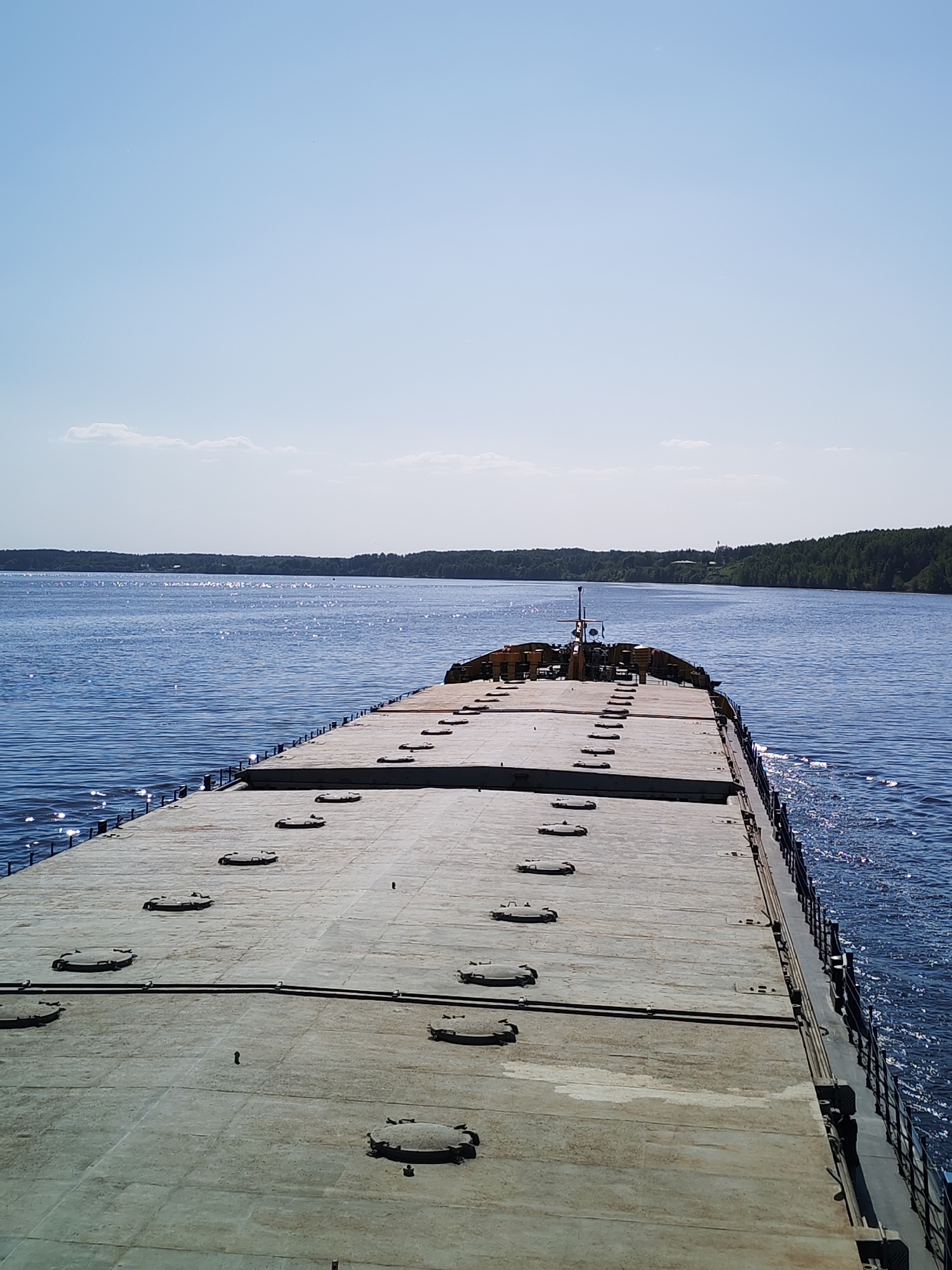 The image shows the deck of a barge or large ship sailing on a body of water. The deck is gray and has numerous circular access hatches. A railing runs along the right edge of the deck. In the distance, land with trees is visible on both sides of the water. The sky is blue with a few scattered clouds. Reflections of sunlight can be seen on the water's surface. The city and location are not discernible from the image.