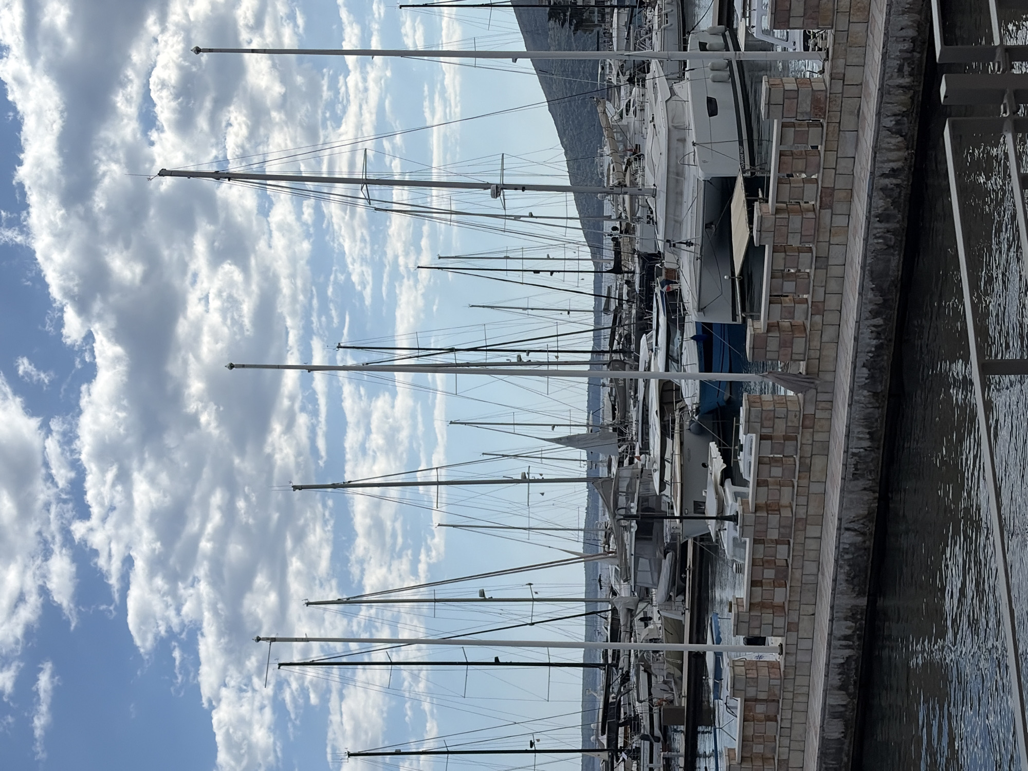The image shows a marina in Meljine, Montenegro. Many sailboats and yachts are docked along a brick pier. The masts of the boats extend high into the sky, which is blue with scattered white clouds. A mountain is visible in the background behind the boats. The water in the marina is dark. A railing is present on the right side of the image.