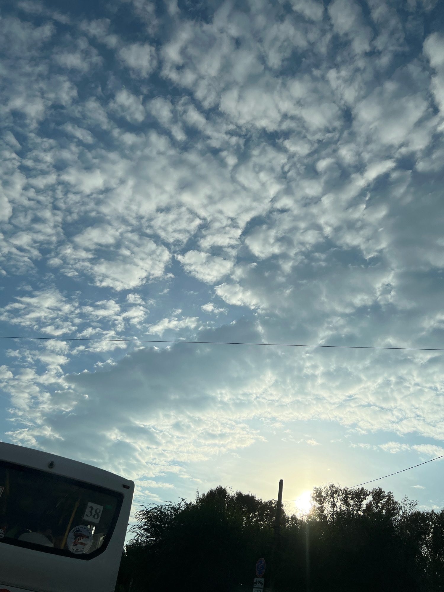 The image depicts a sky filled with scattered clouds and the sun peeking through the foliage of trees. In the lower left corner, a portion of a white bus is visible, showing a window with reflections and a sticker that appears to have a US flag and an eagle. The number "38" is also displayed on the bus window. Silhouetted trees form a dark line across the bottom third of the image, with a pole visible just to the left of the sun. A thin wire stretches horizontally across the sky. The scene suggests an outdoor setting, likely during daytime. There are no identifiable people or explicit actions taking place. The provided information about the location being Krasnoyarsk, Russia, is external to the visual content of the image itself.