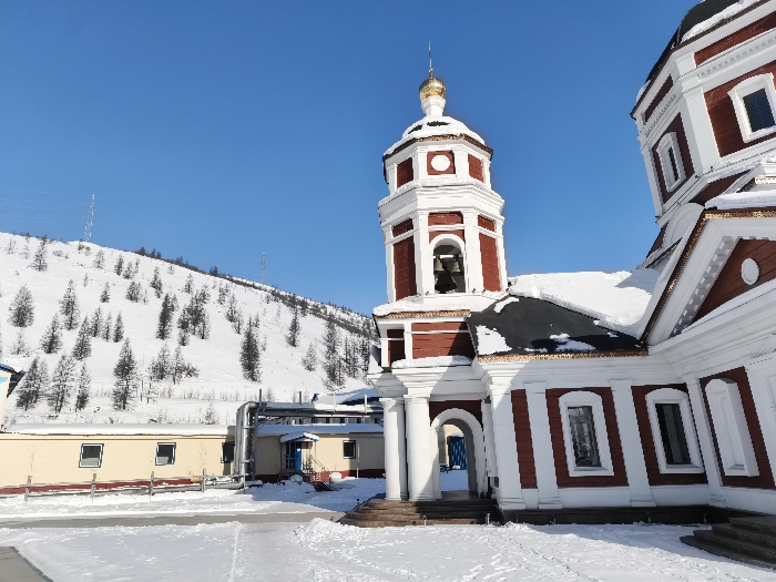 The image displays a winter landscape in the city of Im. Gastello, Russia. A multi-tiered Orthodox church, predominantly red and white with a golden onion dome and spire atop its bell tower, occupies the right side of the frame. The church roofs are heavily covered with white snow, contrasting with its dark roofing material. White architectural elements include columns framing an arched entrance, window surrounds, and cornices. The foreground and visible ground areas are blanketed in undisturbed white snow, with minor impressions suggesting faint pathways. In the background, a large snow-covered hill slopes upwards, sparsely populated by dark, slender trees. Two lattice-style electrical transmission towers are visible on the hillside. Below the hill and to the left of the church, several low-rise, light-colored utility buildings with dark roofs, rectangular windows, and external silver-colored piping are situated. One building features a red outdoor staircase leading to a blue door. A low, light-colored fence is partially visible in front of these utility structures. The sky is clear, bright blue, indicating daylight conditions. No individuals are present in the scene.