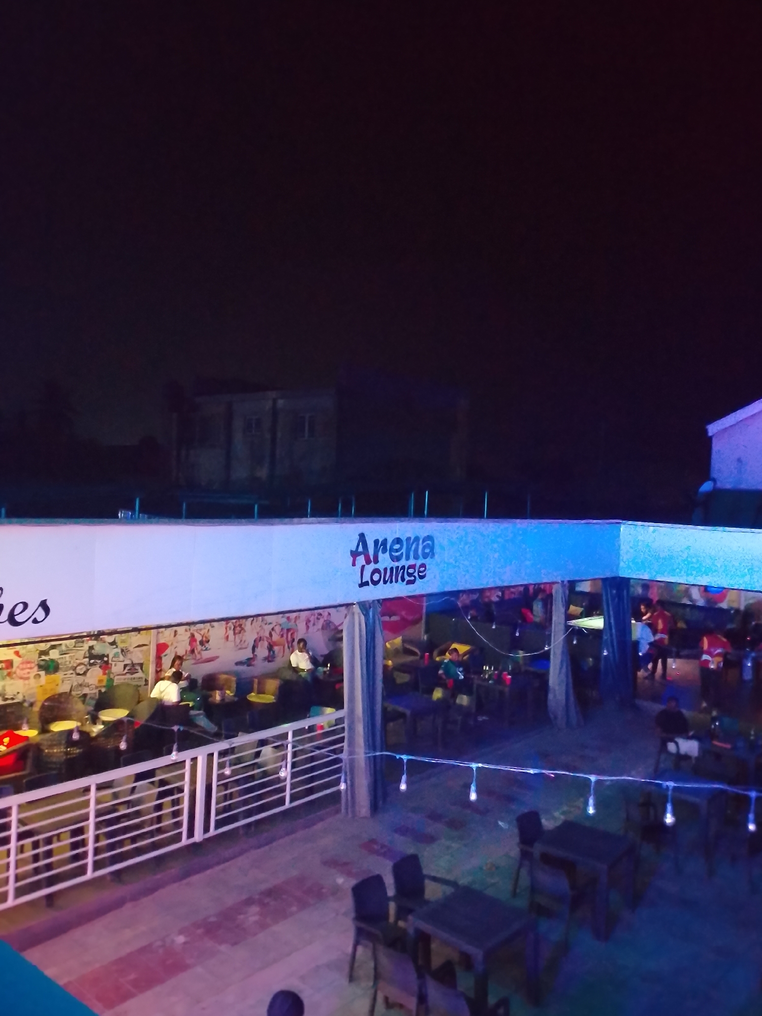 The image shows an outdoor lounge at night in Narayi, Nigeria. The lounge, named "Arena Lounge," is partially covered with a structure bearing its name. Inside the lounge, people are seated at tables, possibly dining or socializing. The lounge is decorated with lights strung across the open area. The flooring of the outdoor area is tiled with a reddish-brown pattern. In the background, there are buildings, and the overall lighting is dim.