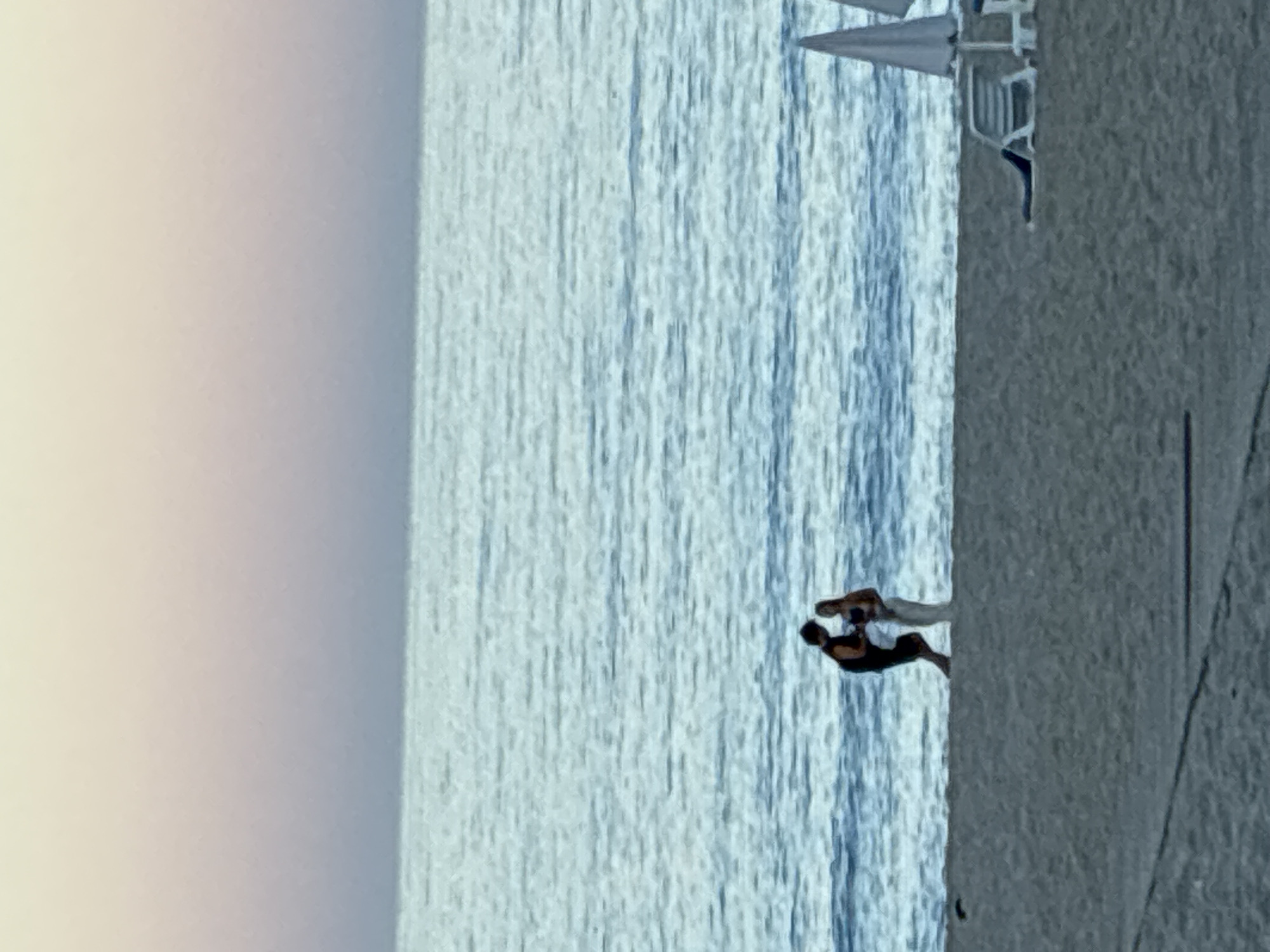 Two people, a man and a woman, stand at the edge of the water on a sandy beach in Alanya, Turkey. The man, wearing a dark tank top and shorts, is on the left. The woman, in a bikini top and light-colored pants, stands to his right. They are facing each other and appear to be interacting. The sea is calm and blue with gentle waves. The sky above is a soft gradient of pink and purple, indicating either sunrise or sunset. In the foreground, the beach is sandy with lounge chairs and umbrellas visible on the right side.