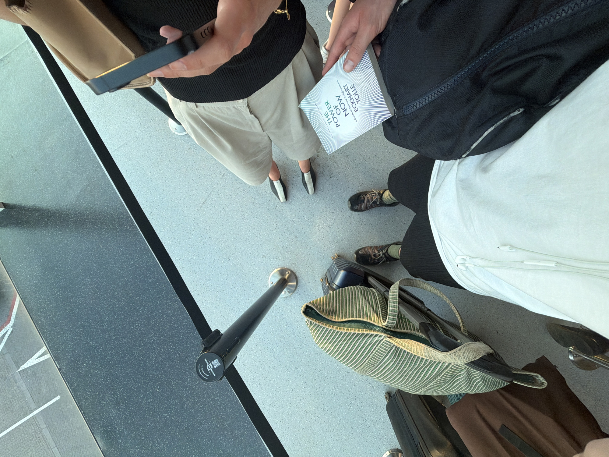 The image shows a group of people in a queue, likely at an airport or train station given the presence of luggage and stanchions. The perspective is from above, looking down at the individuals and their belongings. One person is holding a book titled "THE POWER OF NOW" by Eckhart Tolle. Another person is wearing light-colored shorts and black and white shoes, while another is in black pants and dark shoes. There are several pieces of luggage visible, including a striped duffel bag and a black backpack. The floor is a light gray, speckled surface, and a dark grey barrier or counter runs along the left side of the frame. The lighting suggests it is daytime, possibly indoors with artificial lighting or near a window. The overall mood is neutral, with people waiting. There is some visible text on the book and on a circular tag attached to one of the stanchions, but it is largely illegible.