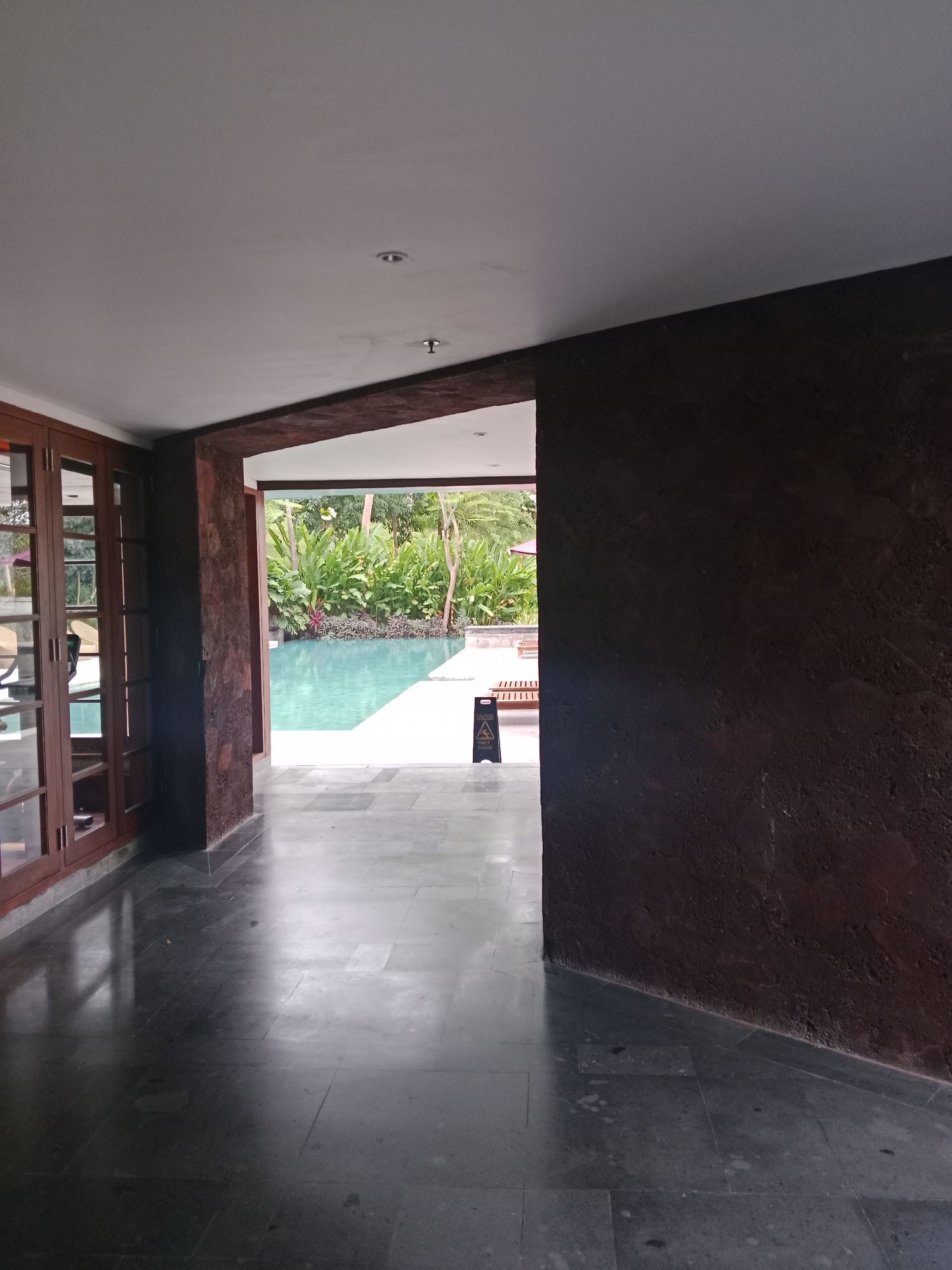 The image shows a hallway with a view of a swimming pool and lush greenery outside. The hallway features dark, polished stone floors and a dark brown textured wall on the right. On the left, there are large wooden-framed glass doors.
In the distance, a clear blue swimming pool is visible, surrounded by vibrant green plants and trees. Sunlight appears to be filtering through the foliage, suggesting it is daytime. A yellow "caution wet floor" sign stands near the edge of the pool. The overall atmosphere is serene and suggests a resort or private residence in a tropical location, consistent with the Kuta Selatan, Indonesia context. There are no people visible in the image.