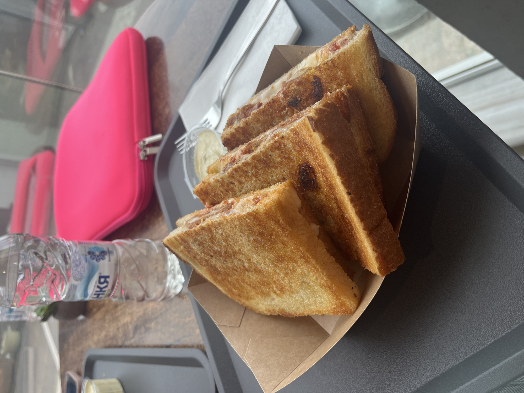 A close-up shot shows a meal on a gray tray. The primary focus is a container of grilled sandwiches. Next to the sandwiches, there is a small container of sauce and a disposable fork. To the left of the tray, there's a bottle of water. Further to the left, a bright pink pouch is also visible on what appears to be a brown surface, likely a table. The scene is indoors, lit by natural light coming from a window visible in the background. The bottle of water appears to have text in Russian which could indicate the location as the city of Sofia, Bulgaria, which has a community of Russian speakers. The overall environment appears to be a casual dining setting, possibly a cafe or fast-food restaurant.