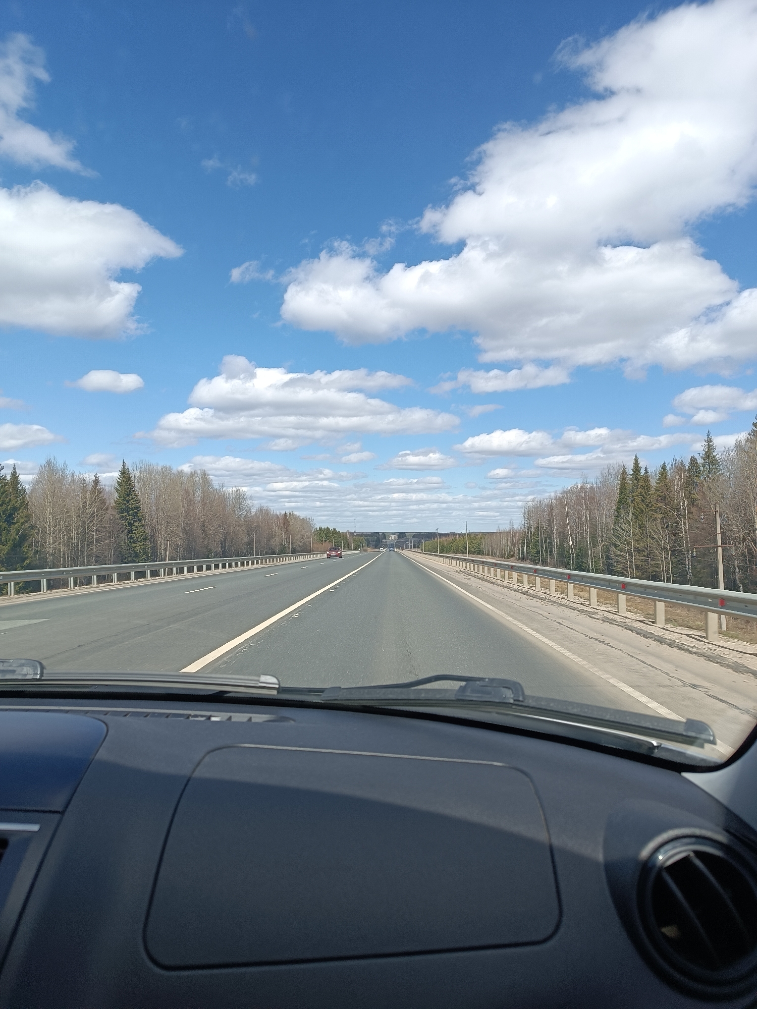 The image depicts a view from inside a car, looking out onto a highway. The dashboard of the car is visible in the foreground. Beyond the windshield is a straight stretch of road with multiple lanes. A dividing line separates the lanes. Guardrails run along the edges of the highway, separating it from forested areas on either side. The trees appear to be a mix of deciduous and evergreen varieties, with many deciduous trees still bare, suggesting it is early spring. In the distance, other vehicles can be seen on the road. The sky is blue with scattered white clouds. The scene suggests travel or commuting on a rural highway. There is no evidence to specifically indicate the location is in the city of Ленинск, Russia.