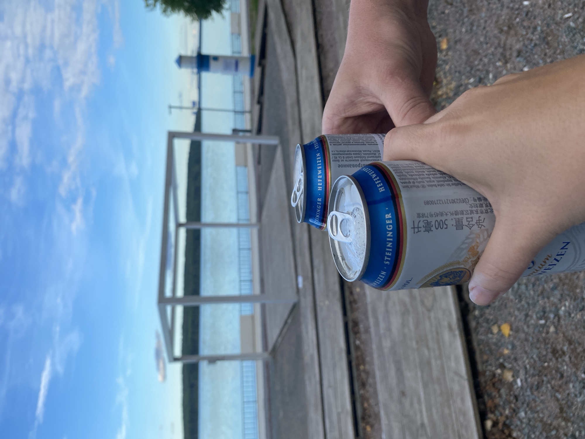 Two hands are holding two opened aluminum beer cans. The cans are blue and white, with "HEFEWEIZEN STEININGER" written on them. In the background, there is a body of water, a wooden platform, and a white structure. Further in the distance, a lighthouse-like structure is visible. The sky is blue with scattered clouds. The image is taken outdoors, likely on a sunny day. The location is the embankment in Chaikovsky, Russia, near the Kama River.