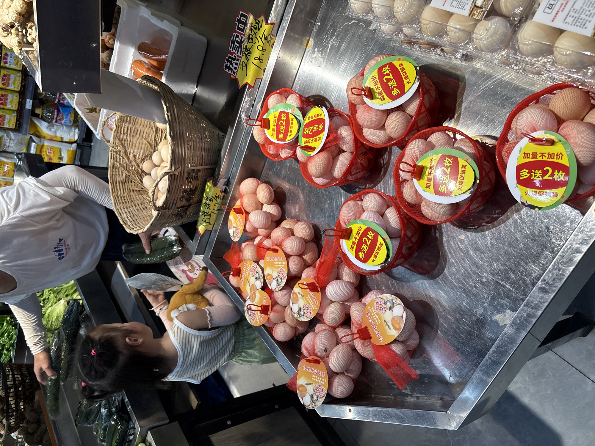 This image depicts a grocery store aisle, specifically in Minhang District, China, with a focus on a display of eggs. A young child, wearing a striped top and light green pants, stands by the display, holding a toy and looking towards a person whose arm is visible reaching for a cucumber. The eggs are packaged in red mesh bags and red plastic baskets, with numerous labels in Chinese. Some of the labels feature images of eggs and text indicating pricing or promotions. In the background, shelves are stocked with various grocery items, including vegetables like cucumbers and lettuce, and bags of dry goods. A woven basket filled with eggs is also visible. The scene appears to be a typical retail environment within a Chinese supermarket.