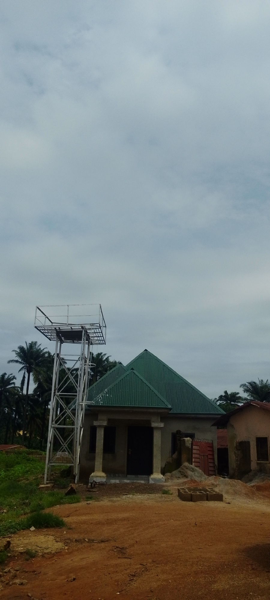 This is a vertical, eye-level shot of an outdoor scene in Baissa, Nigeria. A tall, metallic water tower stands on the left of the frame, with a square platform at its apex. To the right of the water tower is a building with a green, corrugated metal roof that has a prominent, pointed apex. The building appears to be under construction, with rough concrete pillars framing a dark doorway. A pile of bricks and sand sits to the right of this building. Further to the right, a smaller, lighter-colored building with a reddish-brown roof is visible. The foreground is a reddish-brown dirt path with scattered rocks and some square concrete blocks. Lush green grass and palm trees are visible to the left and behind the structures. The sky is overcast with diffused light.